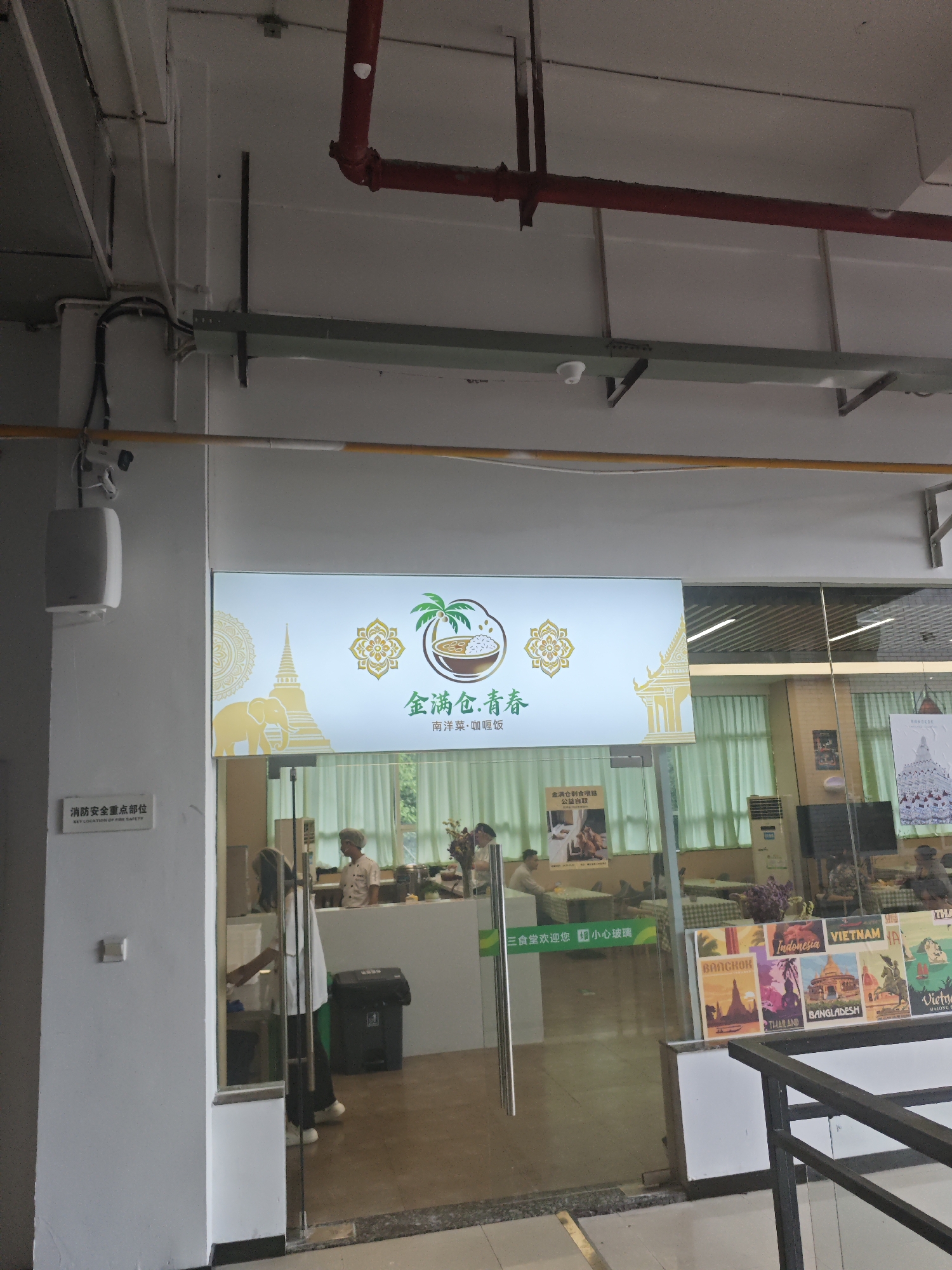 An interior scene at what appears to be a restaurant or food service area within the Chongqing High-tech Zone, China. The main subject is a backlit sign displaying Asian-themed graphics, including an elephant, a pagoda, and a bowl of food with a palm tree. The sign has Chinese text which can be partially translated as "Golden Full Bowl, Youthful". Below this, in smaller characters, it reads "South Ocean Food, Coffee, Rice".
To the left of the sign, a white wall is visible with a mounted white speaker and a security camera. A small sign on the wall reads "Key Location of Fire Safety" in both Chinese and English.
The foreground features a glass entrance with people visible inside. Through the glass, a counter area is seen with employees in food service attire, one wearing a chef's hat. Several tables with customers are also visible in the background, along with a large air conditioning unit and decorative posters on the right side of the glass entrance. These posters depict travel destinations like Bangkok, Thailand, Indonesia, and Vietnam.
The overall environment suggests a public indoor space, likely a food establishment, with operational staff and patrons. The lighting appears to be a mix of overhead artificial lighting and the illumination from the prominent backlit sign.