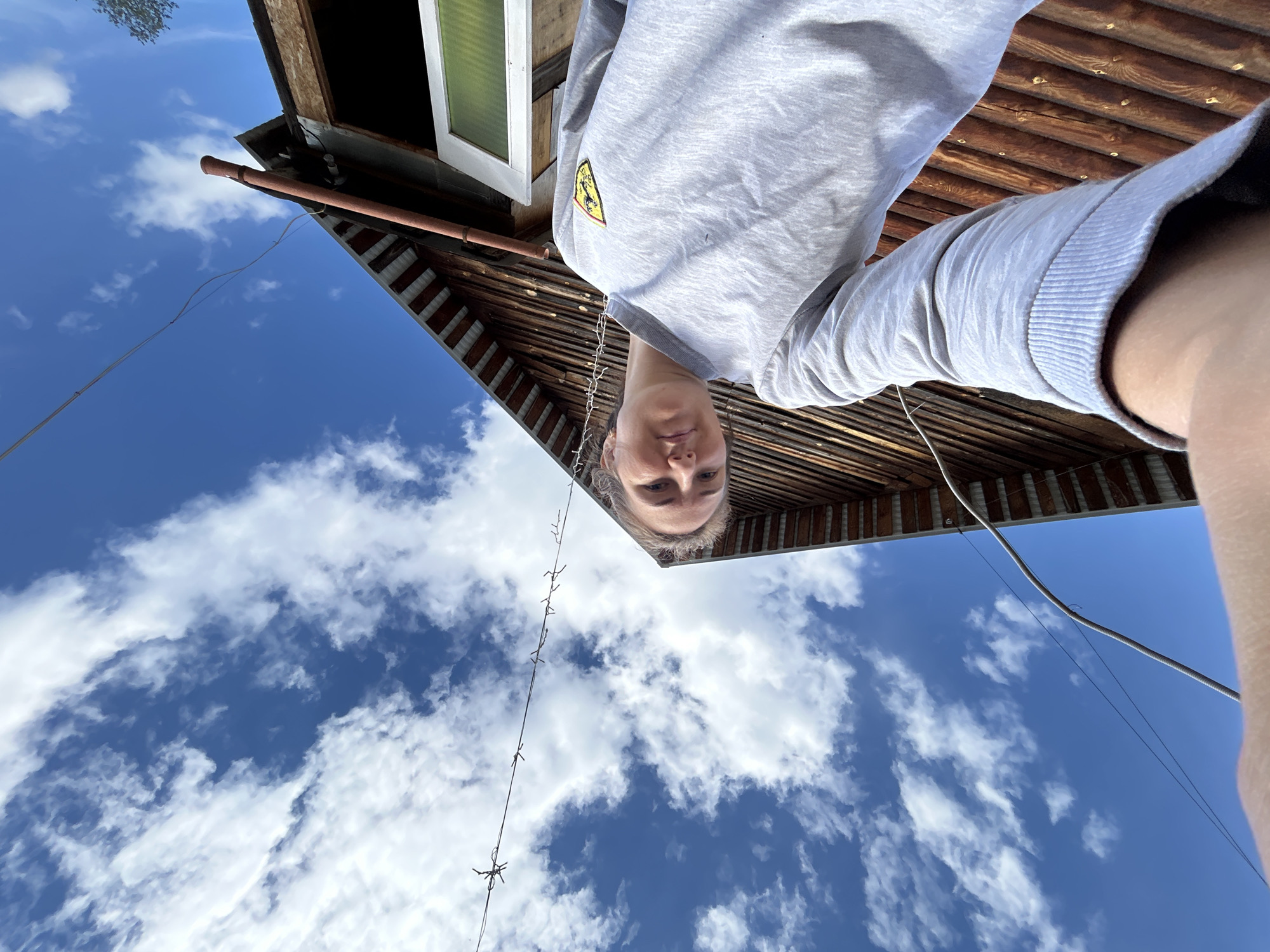 A low-angle photograph depicts a woman in a grey sweatshirt, positioned against a backdrop of a bright blue sky dotted with white clouds, and a rustic wooden building with a white-framed window. The woman is looking down at the camera. A thin cable with evenly spaced attachments extends from the building towards the bottom of the frame. The location is Noviy, Russia.