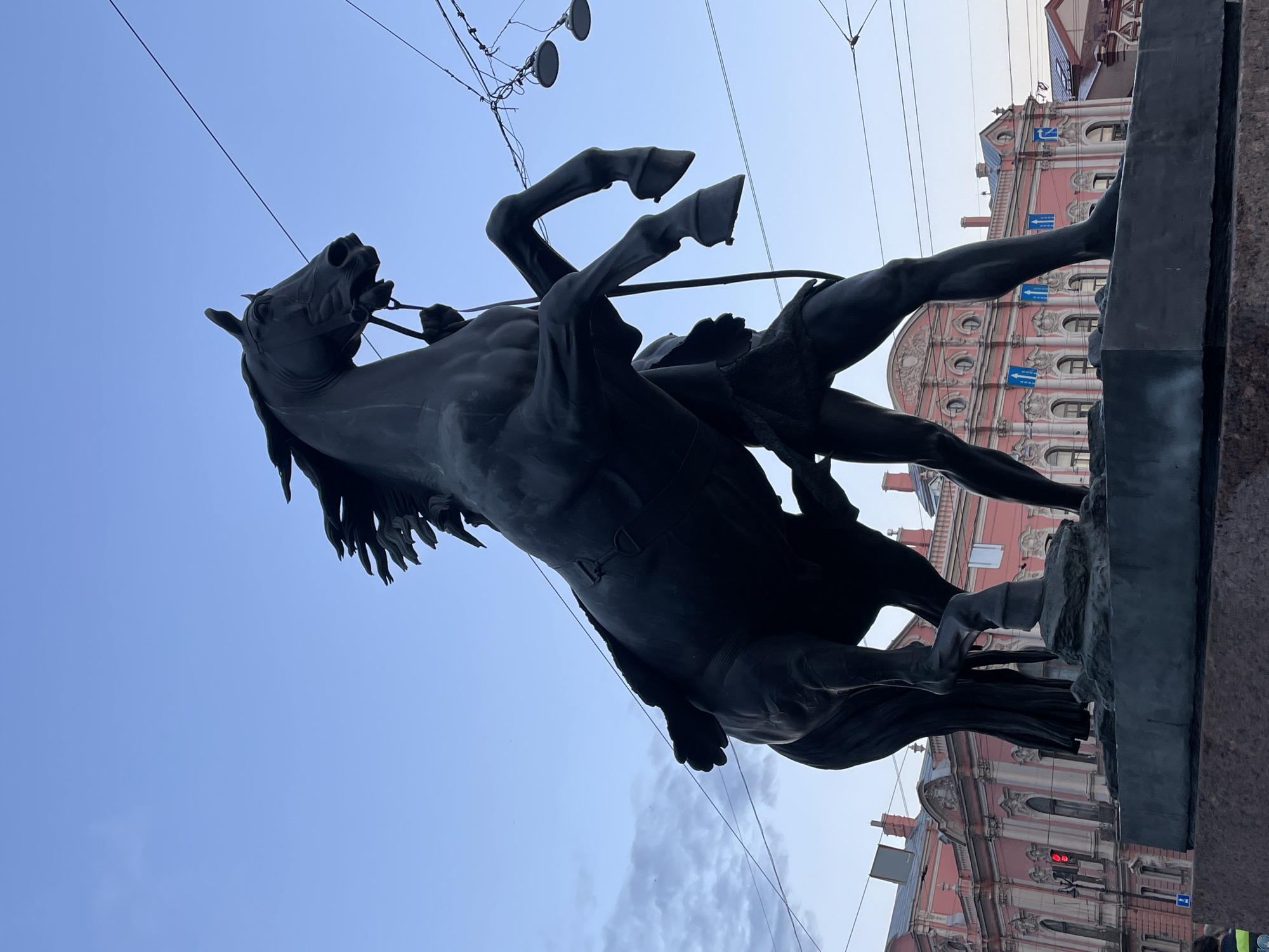 The image shows the Bronze Horseman monument in St. Petersburg, Russia, a bronze equestrian statue of Peter the Great. The statue is dark and silhouetted against a pale blue sky with some clouds on the left. The horse is rearing up on its hind legs, with its front legs in the air, and a figure, presumably Peter the Great, is mounted on it, holding the reins. The monument stands on a large stone pedestal. In the background, there is a light pink building with classical architecture, featuring multiple windows and decorative elements. There are also overhead wires and streetlights visible. The scene appears to be in a public square or street.