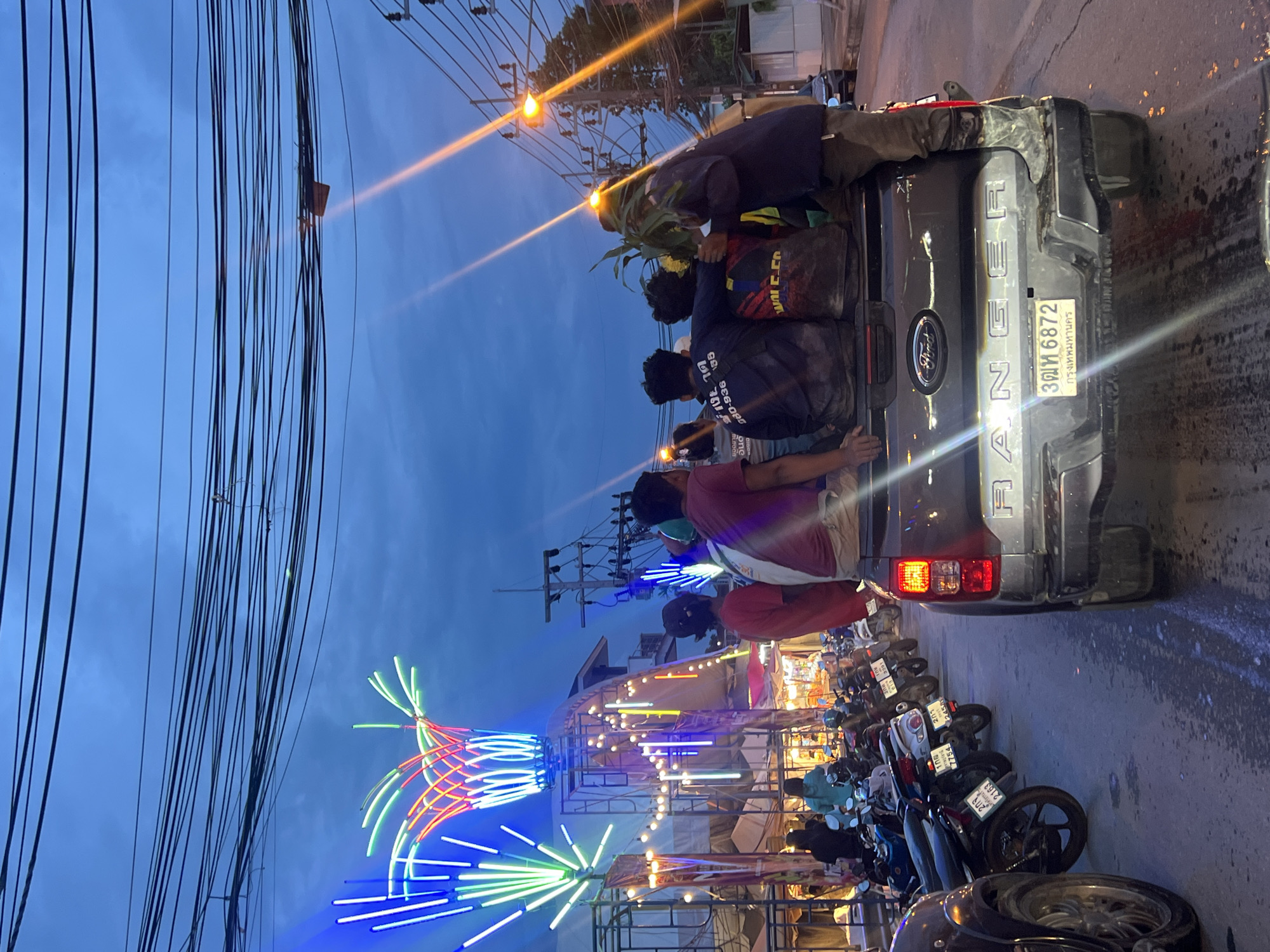 Here is a detailed description of the image:
**General Description:** The image appears to be taken in a street scene at dusk or early evening. The focus is on the back of a dark gray Ford Ranger pickup truck, with several people standing in the bed of the truck. The street seems to be lined with parked motorbikes and there are light displays and electrical wires overhead.
**Specific Details:**
* **People:** At least four individuals are in the bed of the truck, but their faces are not very clear. They seem to be standing and possibly working with something, as one person is holding green plants.
* **Vehicles:** The main subject is the Ford Ranger pickup truck. Several parked motorbikes line the street in front of the truck.
* **Light Displays:** There are elaborate light displays with neon-colored tubes (blue, green, red) that resemble festive decorations.
* **Environment:** The scene is an urban area, with visible electrical wiring and utility poles overhead.
* **License Plate:** The license plate of the truck reads "32ห 6872". The license plate also contains Thai text.
* **Objects:** In addition to the vehicles, lights, and wires, there are several miscellaneous objects visible, including some bags in the bed of the truck.
**Activities:** The people in the truck appear to be involved in some kind of activity possibly related to setting up or taking down the light displays or other decorations.
**Location:** It is not possible to tell the specific city.