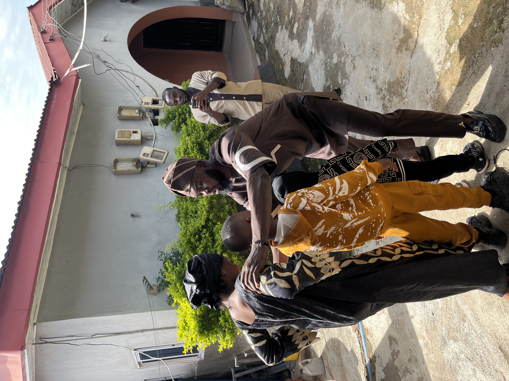In Jos, Nigeria, under a cloudy sky, a man in a brown kaftan and kufi approaches a young boy in a yellow tracksuit, placing a hand on the boy's shoulder. The boy, facing away from the camera, is being guided by a woman in a black dress with gold embroidery, who stands between the man and the boy. A second man, dressed in a light brown and tan outfit, stands in the background, looking at his mobile phone. The setting appears to be the exterior of a residential building with a grey stucco finish, a red corrugated metal roof, and an arched doorway. Green foliage is visible behind the group, and the ground is a concrete surface with some debris. Electrical conduits and utility boxes are mounted on the wall of the building.