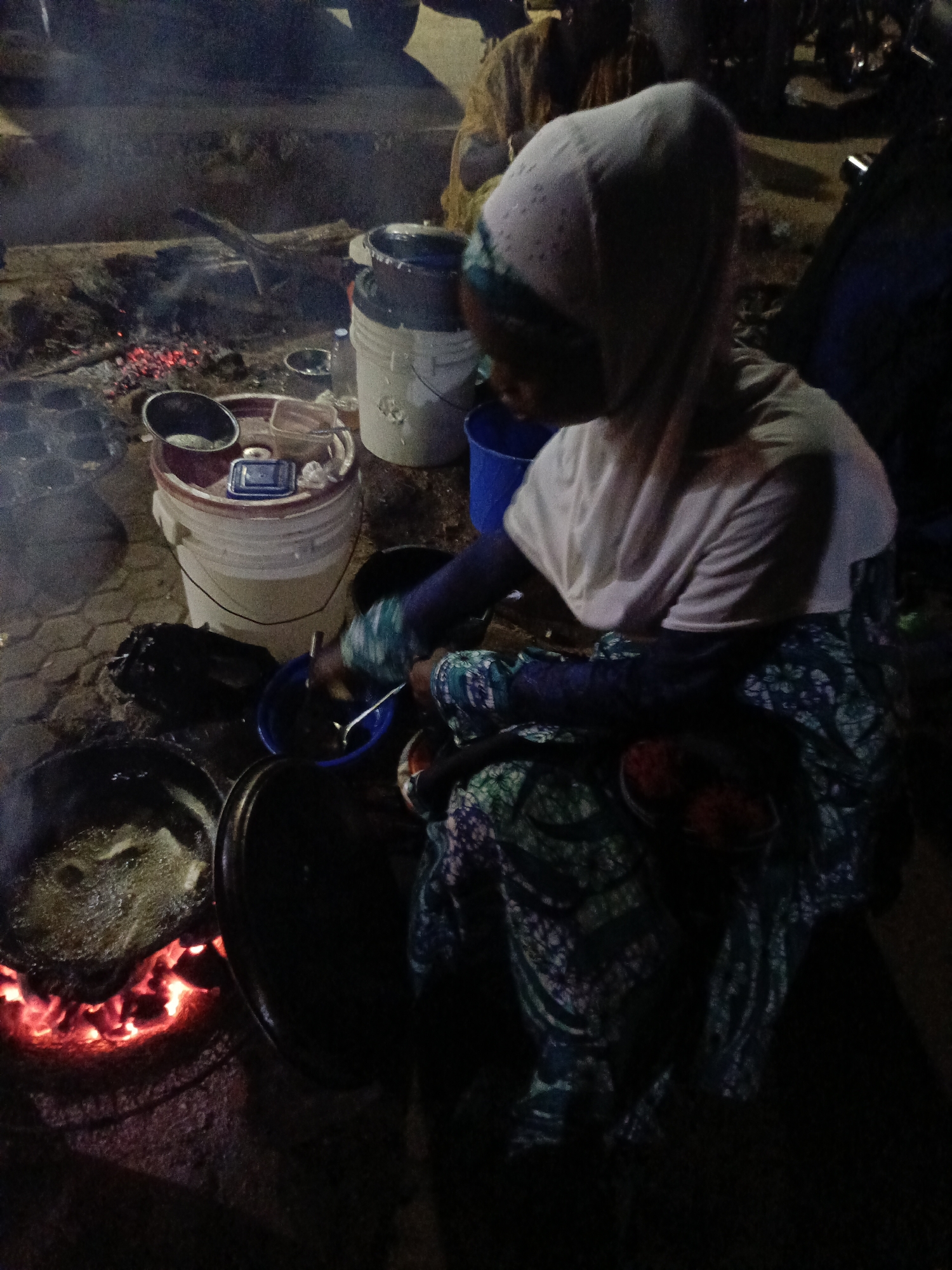 In Akwanga, Nigeria, a woman wearing a headscarf and patterned clothing sits near a fire. She appears to be cooking food in a large pan over the fire. There are buckets and other containers nearby, along with a small pile of burning wood.
