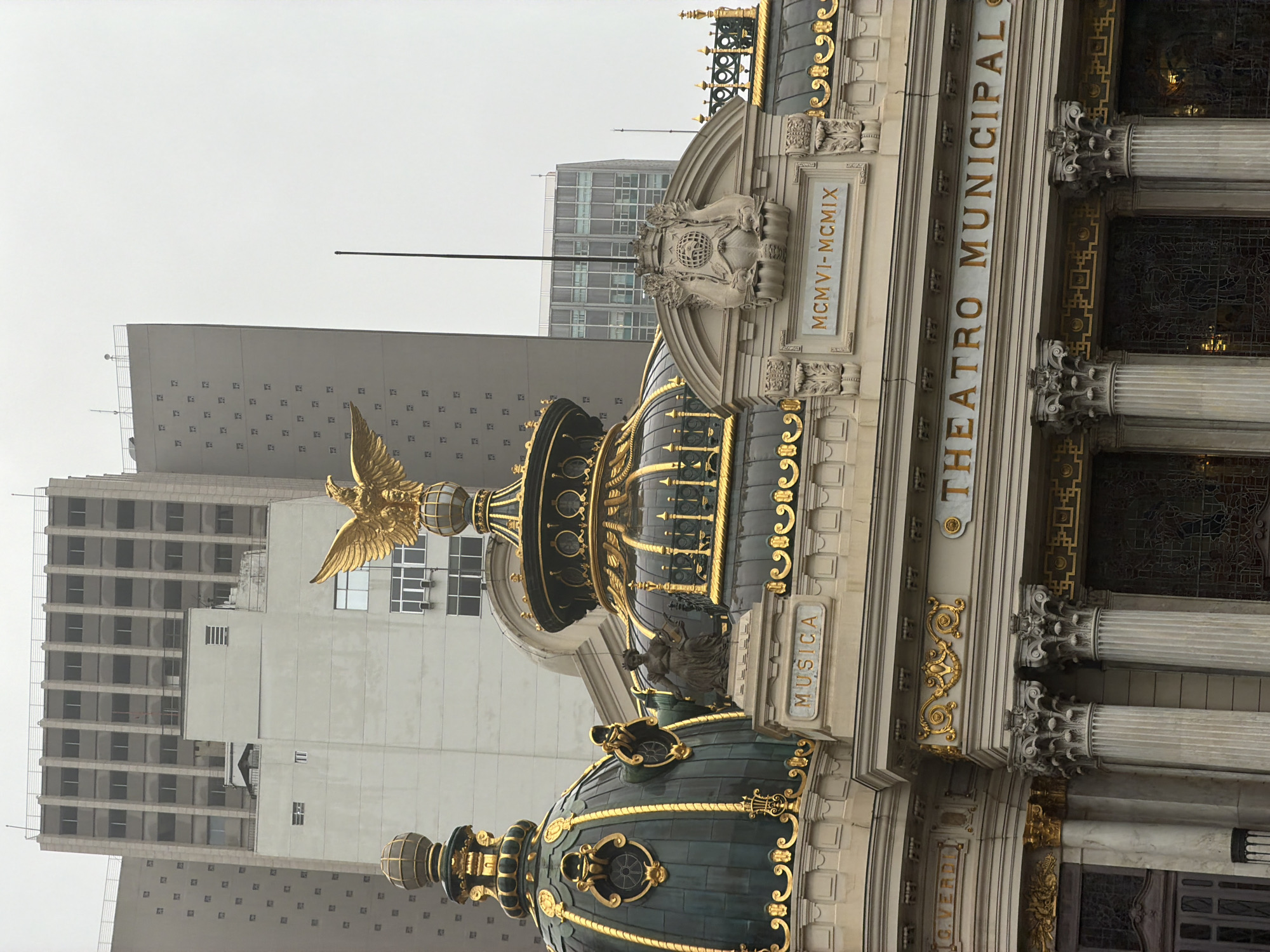 The image shows a close-up view of the facade of the Theatro Municipal in Rio de Janeiro, Brazil. The building features ornate architectural details, including gilded domes, classical columns, and decorative sculptures. A prominent golden eagle sculpture is visible atop one of the domes. Inscriptions on the facade include "MUSICA" and "THEATRO MUNICIPAL", as well as Roman numerals "MCMVI-MCMIX". Several modern, tall buildings rise behind the historic theater, suggesting an urban setting. The sky is overcast, and the overall lighting is diffused. No people are visible in the image.