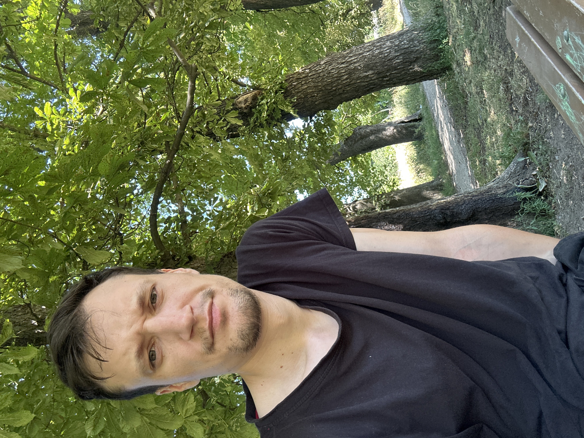 The image shows a man with short dark hair and a light beard, wearing a black t-shirt. He is sitting outdoors under a large tree with green leaves, possibly in a park. The background shows a dirt path and more trees. The location is specified as Makiv, Ukraine.