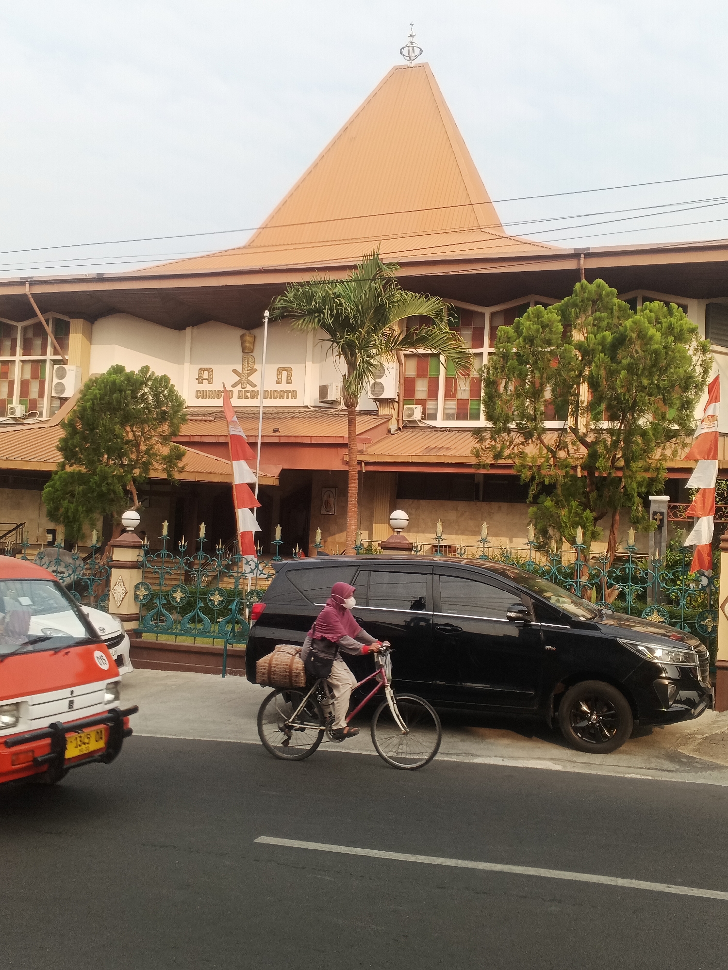 In Sokanegara, Indonesia, a person wearing a pink hijab and face mask rides a bicycle laden with a large woven basket. The bicycle has a red frame and a white front wheel, while the rear wheel appears black. The person is cycling on a paved road alongside a black SUV. In the background, a building with an orange, peaked roof features the inscription "AXN CHRISTO REGICATA" above the entrance. Two Indonesian flags, red and white, are planted near the building. An orange and white van is parked to the left of the SUV. The sky is overcast.