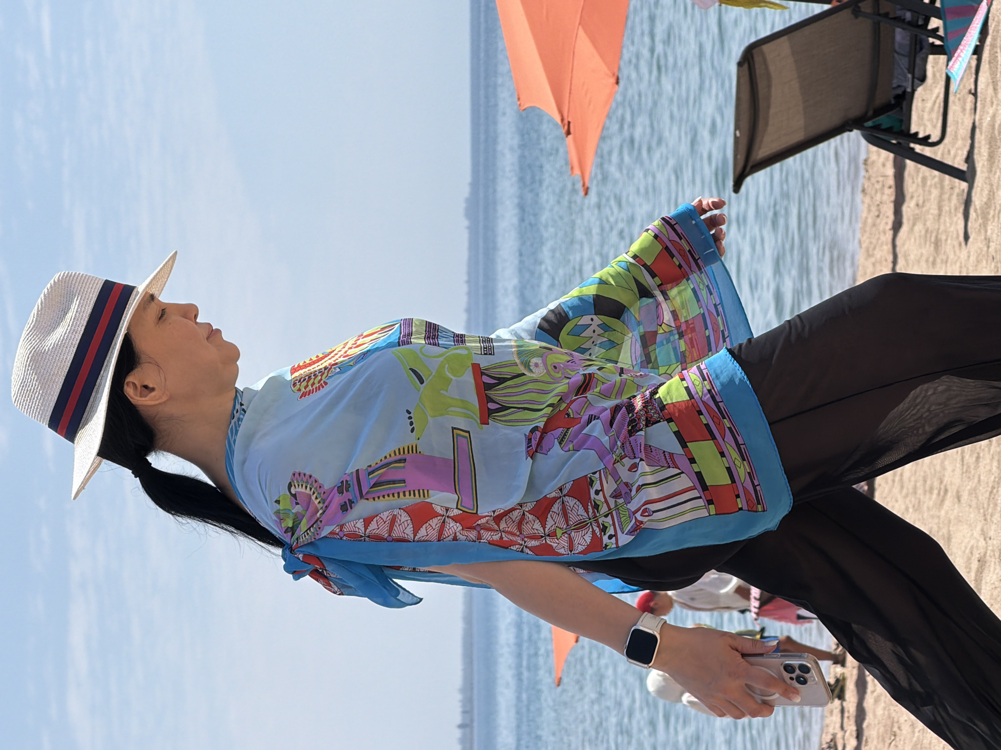 A person wearing a wide-brimmed hat, a light blue patterned tunic, and black sheer pants walks along a sandy beach next to a body of water. The tunic features a brightly colored print with figures and geometric shapes. The person is holding a smartphone in their right hand and wears a white smartwatch on their left wrist. In the background, an orange beach umbrella and a lounge chair are visible. The sky is clear and blue. The location is unknown.
