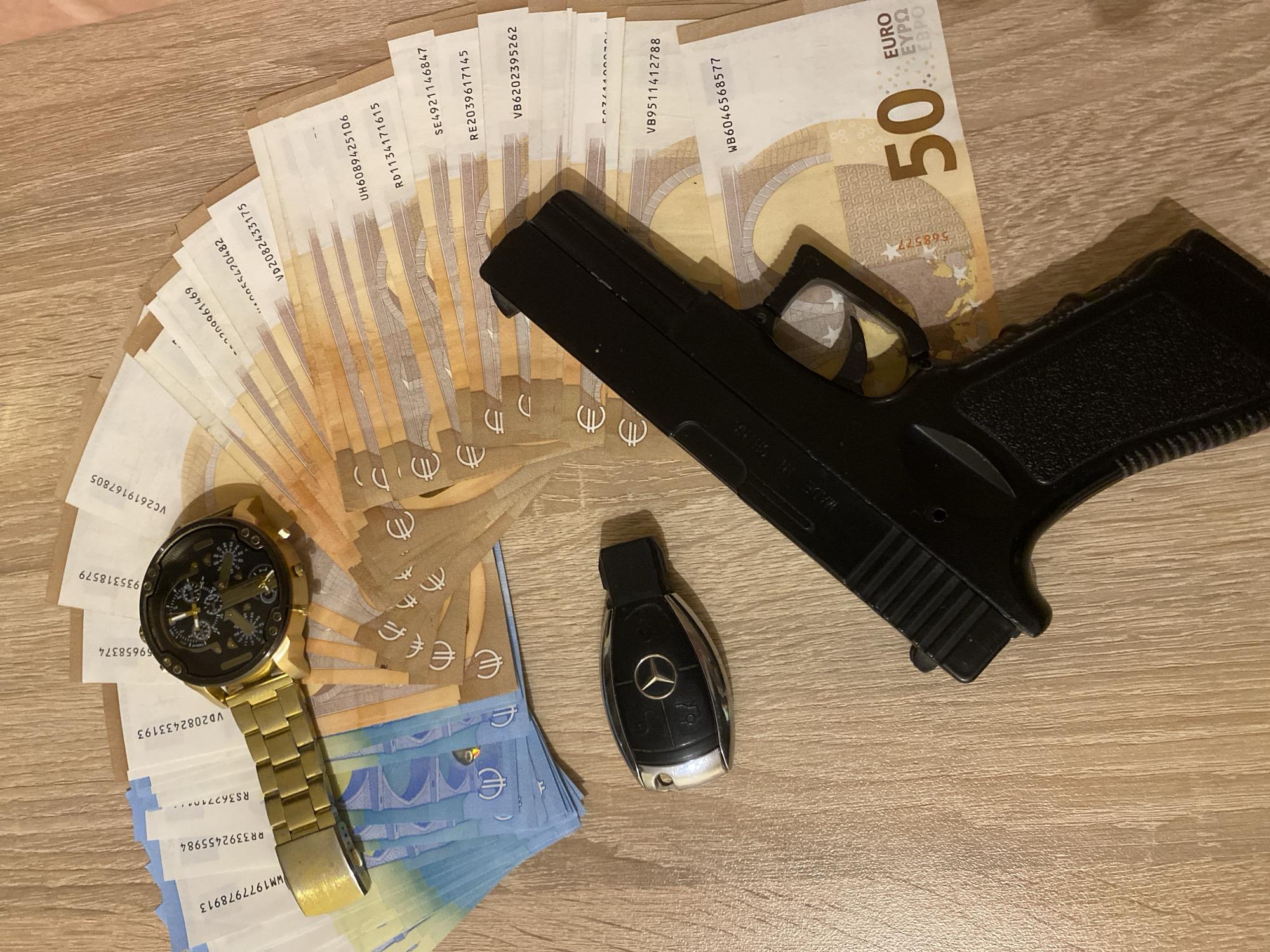 The image shows a collection of luxury items laid out on a wooden surface. There is a large wad of euro banknotes, predominantly 50 euro bills, arranged in a fanned-out manner. A gold watch with a complex dial design is positioned on top of the bills. Adjacent to the money and watch, there is a black toy gun. Additionally, a car key, recognizable as a Mercedes-Benz key, is placed near the gun. The scene is brightly lit, showcasing the details of each item.