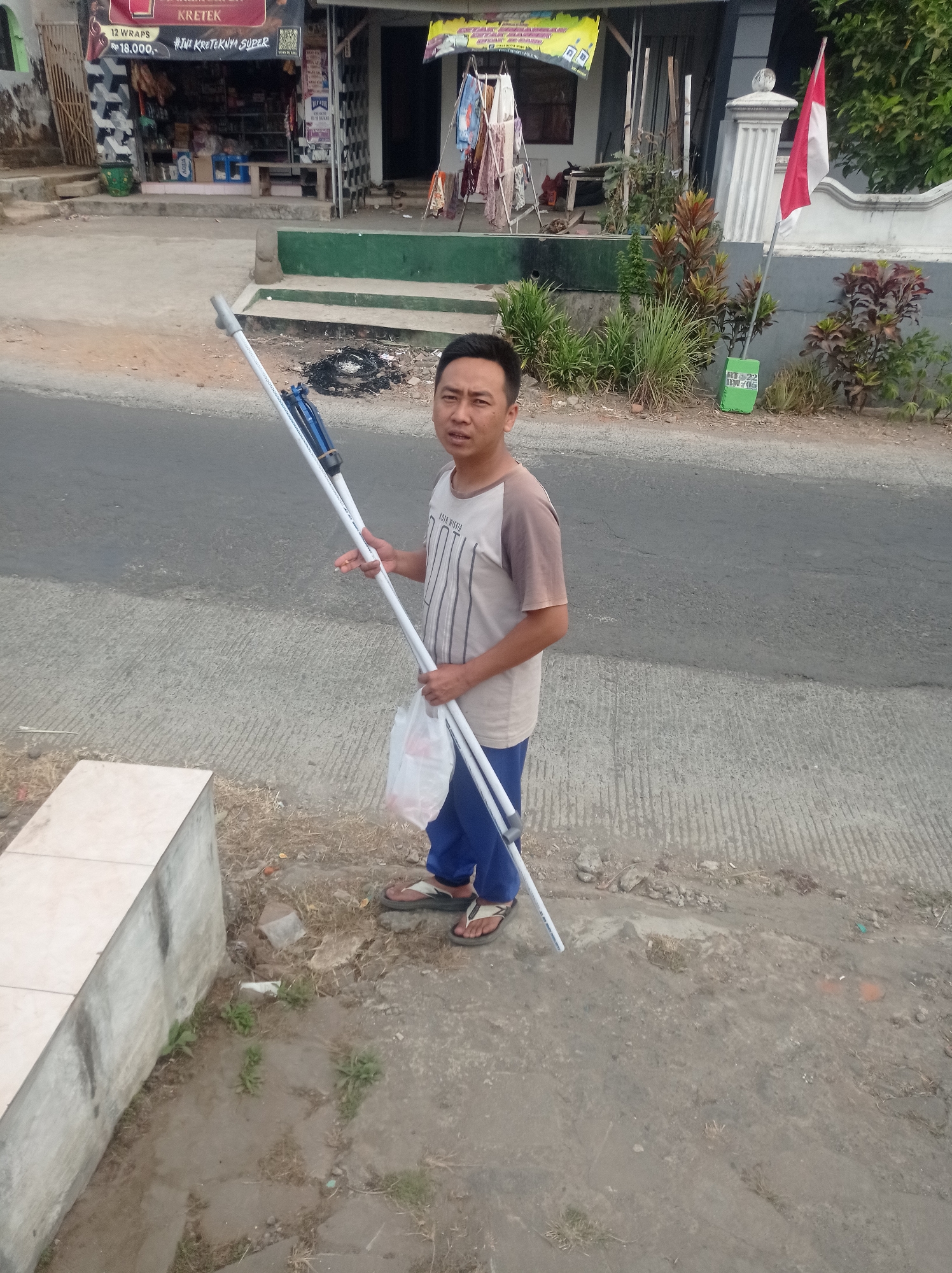 A man stands on the side of a street in Purwosekar, Indonesia, holding a long, white pole with blue and silver components. He is wearing a light brown t-shirt with a dark pattern and blue pants, and flip-flops. He is carrying a white plastic bag in his left hand. The street appears to be paved and there is a concrete structure to the left of the man. Behind him is a building with a storefront, featuring various items displayed for sale, including what appears to be cigarettes and clothing. A red and white Indonesian flag is visible on a flagpole near the building. The environment suggests a commercial area.