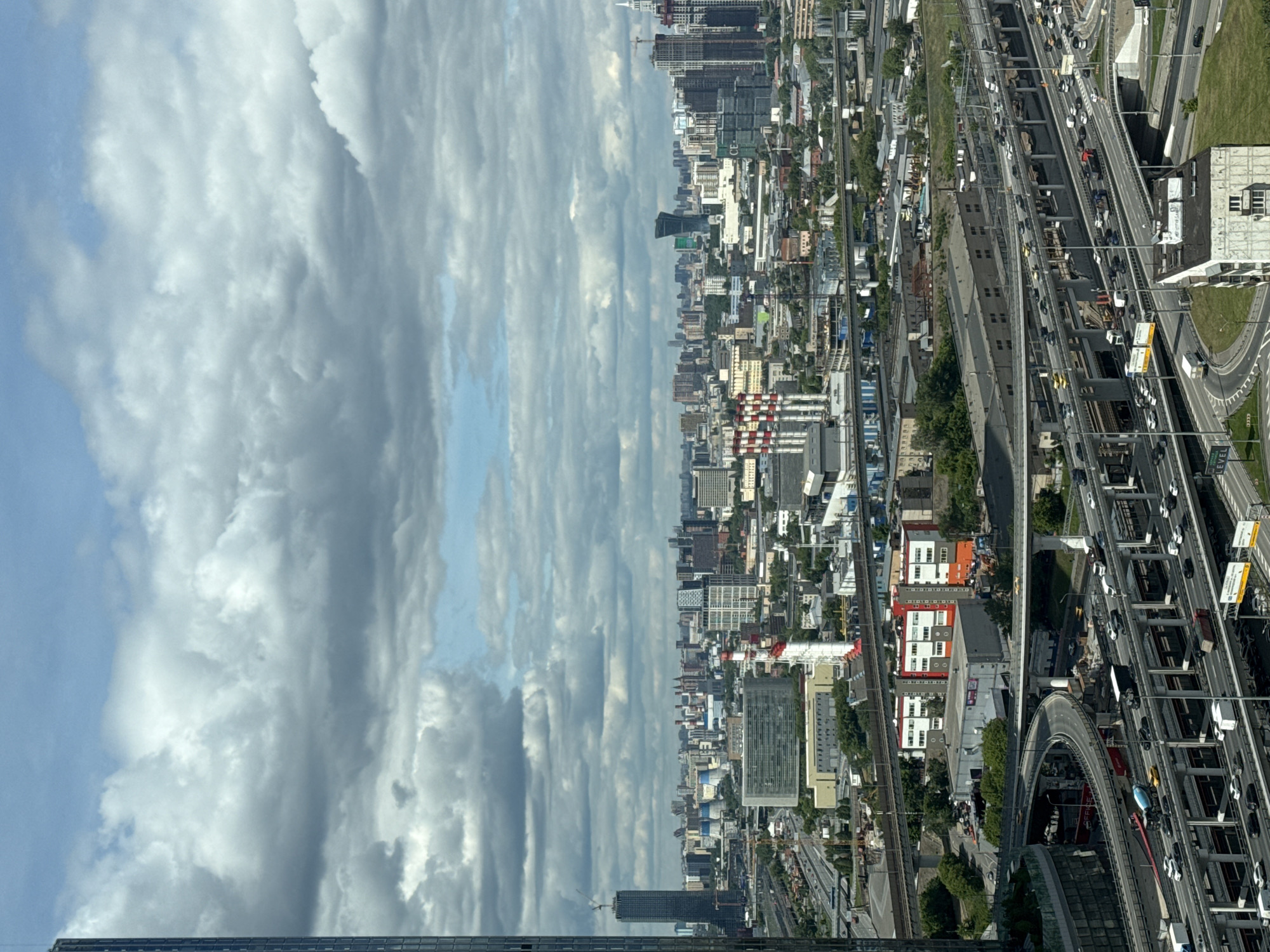 The image shows an aerial view of Khimki, Russia. The skyline is visible with a mix of high-rise buildings and residential structures. A complex highway interchange and multiple lanes of traffic fill the bottom right of the frame. A railway runs parallel to the highway. The left side of the image is dominated by cloudy skies.