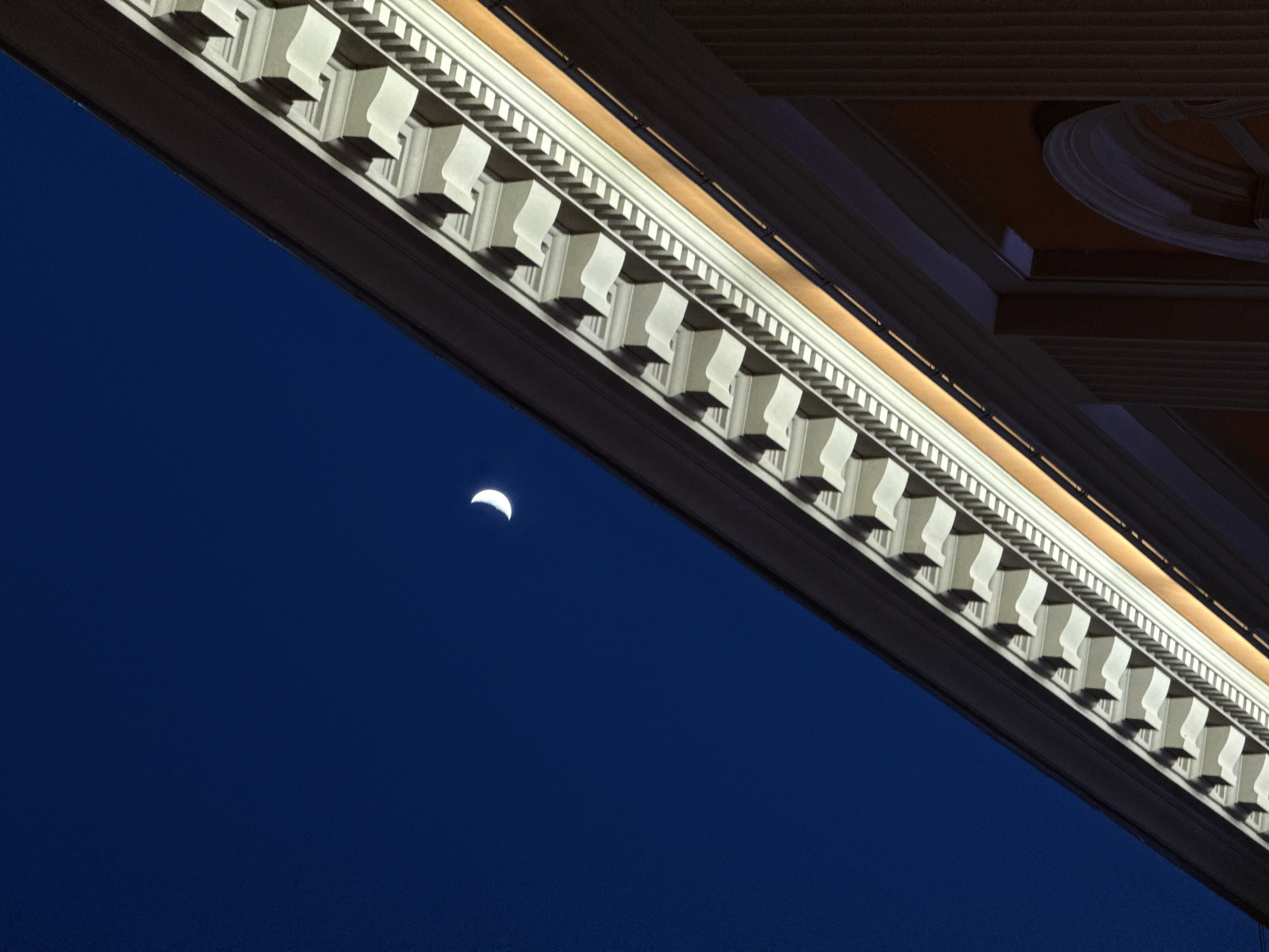 The image captures a crescent moon in a dark blue sky, framed by an ornate, illuminated building structure. The building features a decorative, white facade with a repeating pattern of rectangular and curved elements along its edge. The setting suggests an urban environment at night. Given the architectural style, the image may potentially be located in a European city. If I were to speculate about the city of Moscow, Russia I would also need additional information to verify it.