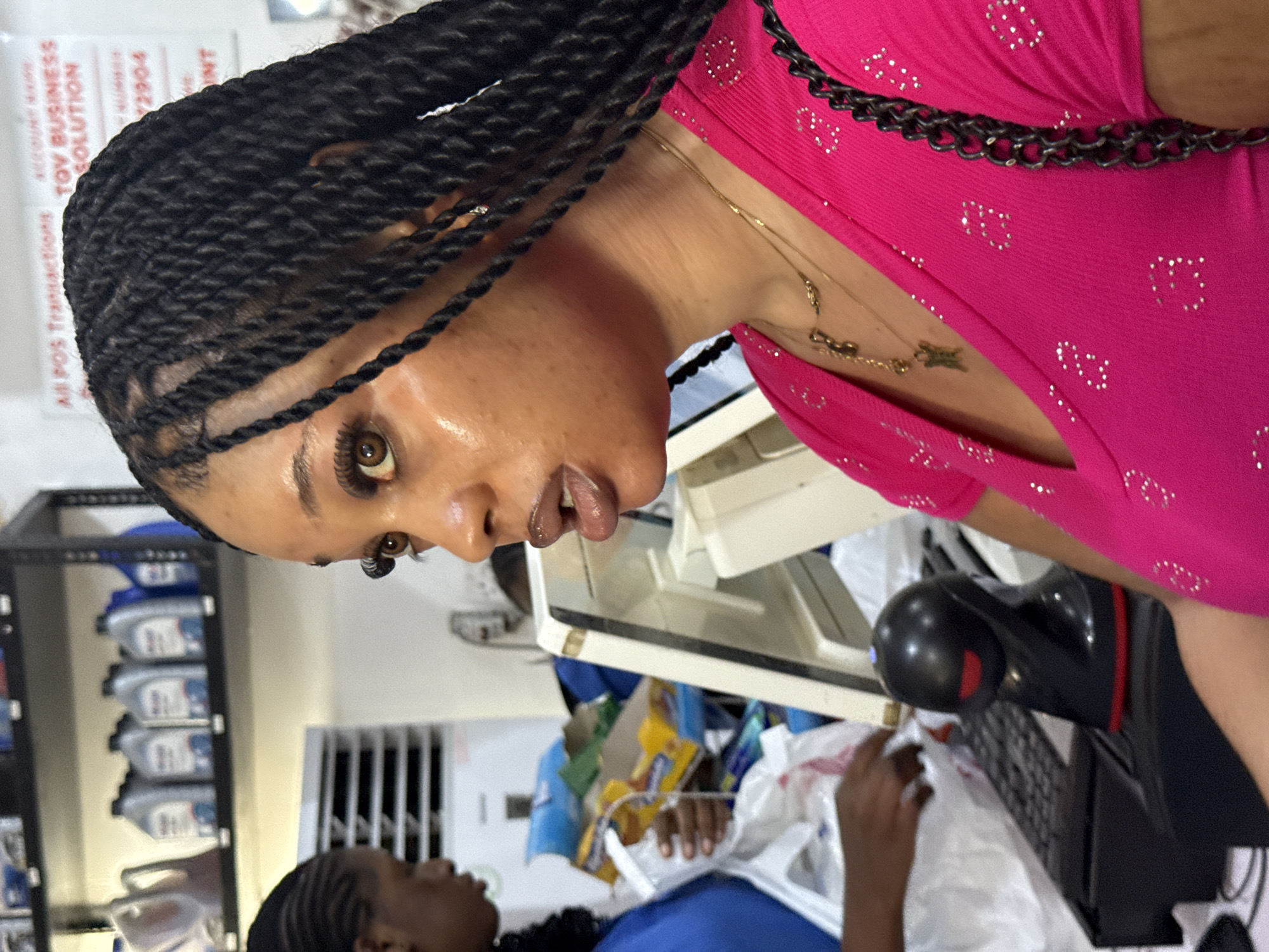The image depicts two dark-skinned women with braided hairstyles in an indoor environment. The woman in the foreground is wearing a bright pink top with what appears to be a black chain detail on the neckline. She is also wearing a gold necklace. The other woman is wearing blue. They are in what seems to be a business setting, near a table with various items, including a keyboard and a monitor, and possibly receipts. Bottles of automotive oil are on shelves in the background. Based on the signs that are partly visible, it could be an "All POS Transactions" place. The location is possibly Ado, Nigeria, based on the prompt.
