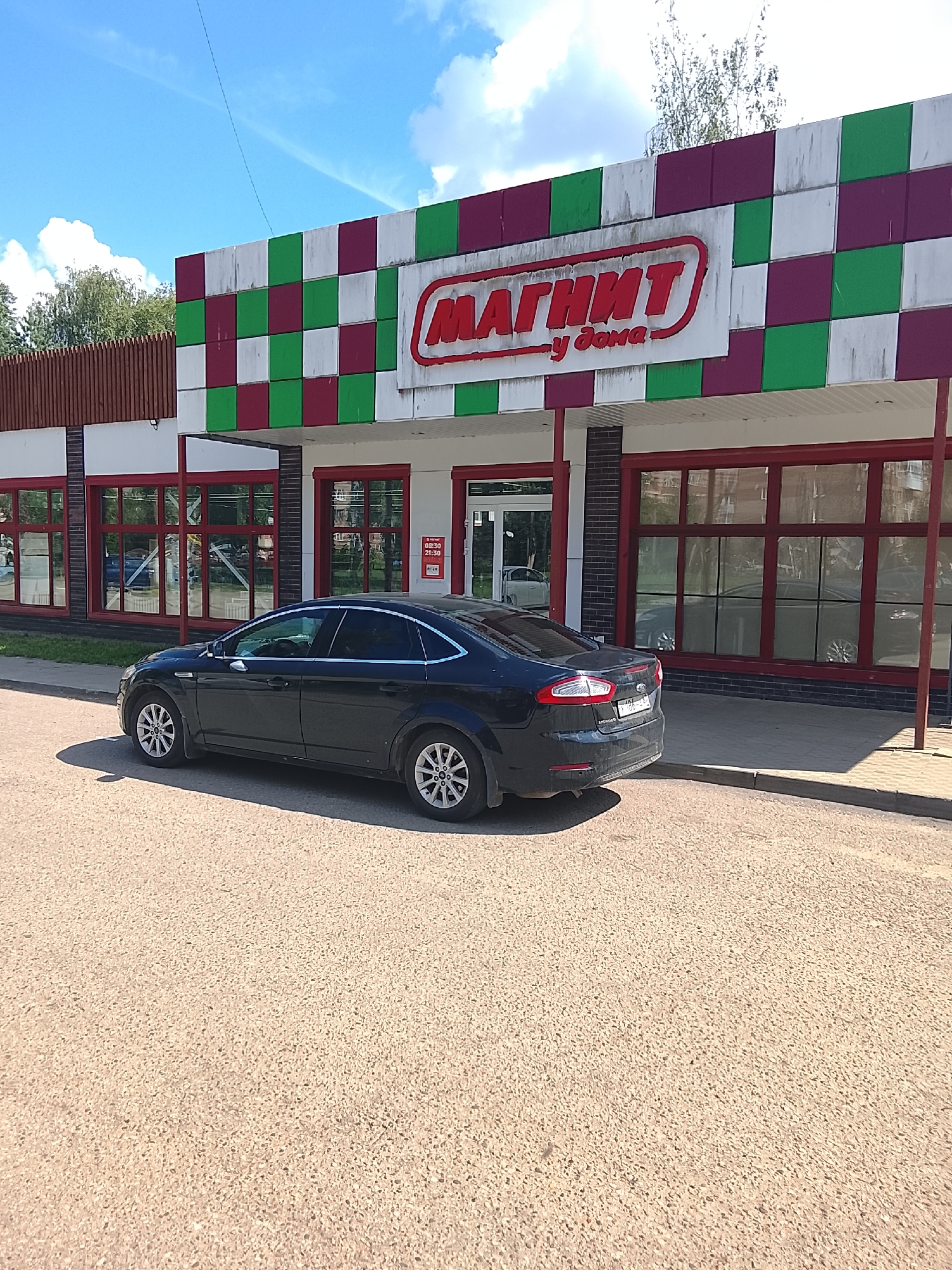 A black Ford Mondeo sedan is parked on a paved road in front of a "Magnit" grocery store. The store has a distinctive facade with a checkerboard pattern of red, green, and white tiles. Large windows with red frames line the front of the store, reflecting the sky and surrounding trees. A sign above the entrance reads "МАГНИТ у дома" in red lettering. The sky is blue with some white clouds. The ground is asphalt with some gravel visible.