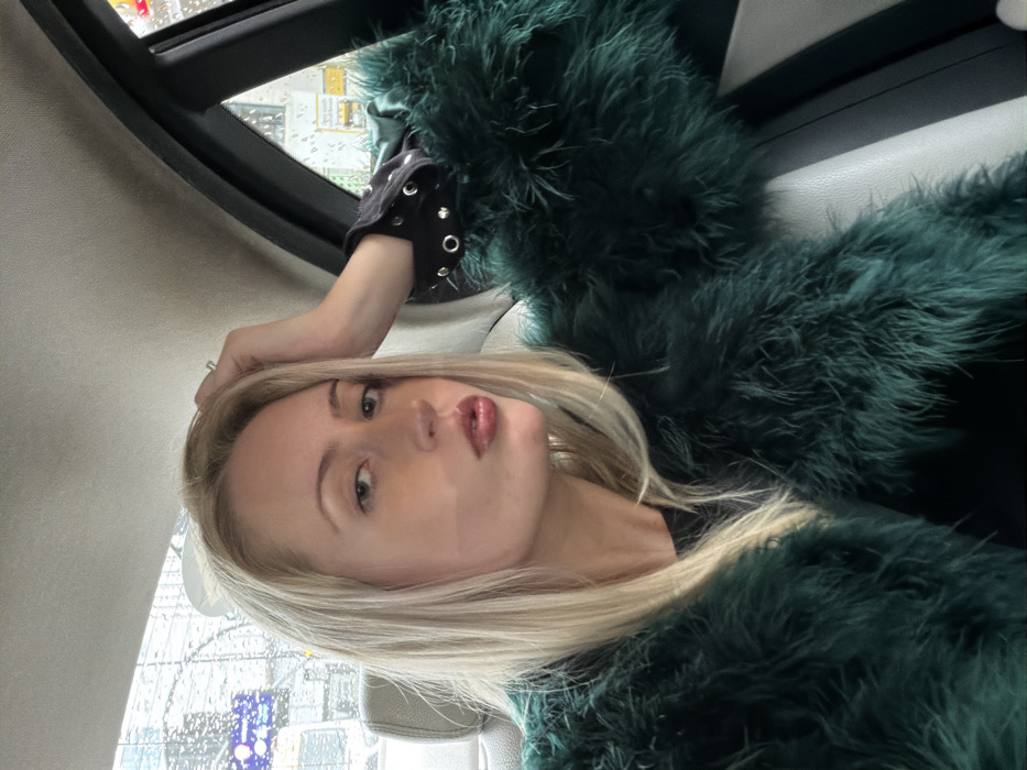 A woman with blonde hair poses in what appears to be a car. She is wearing a dark green, feathered coat or garment. Her left hand is near her head, with a ring visible on her finger. Through the car window, it seems to be a rainy day, with raindrops distorting the view of buildings and streetlights. She has light skin, prominent cheekbones, and her lips are painted with a glossy color. She is wearing a black shirt with metallic studs.