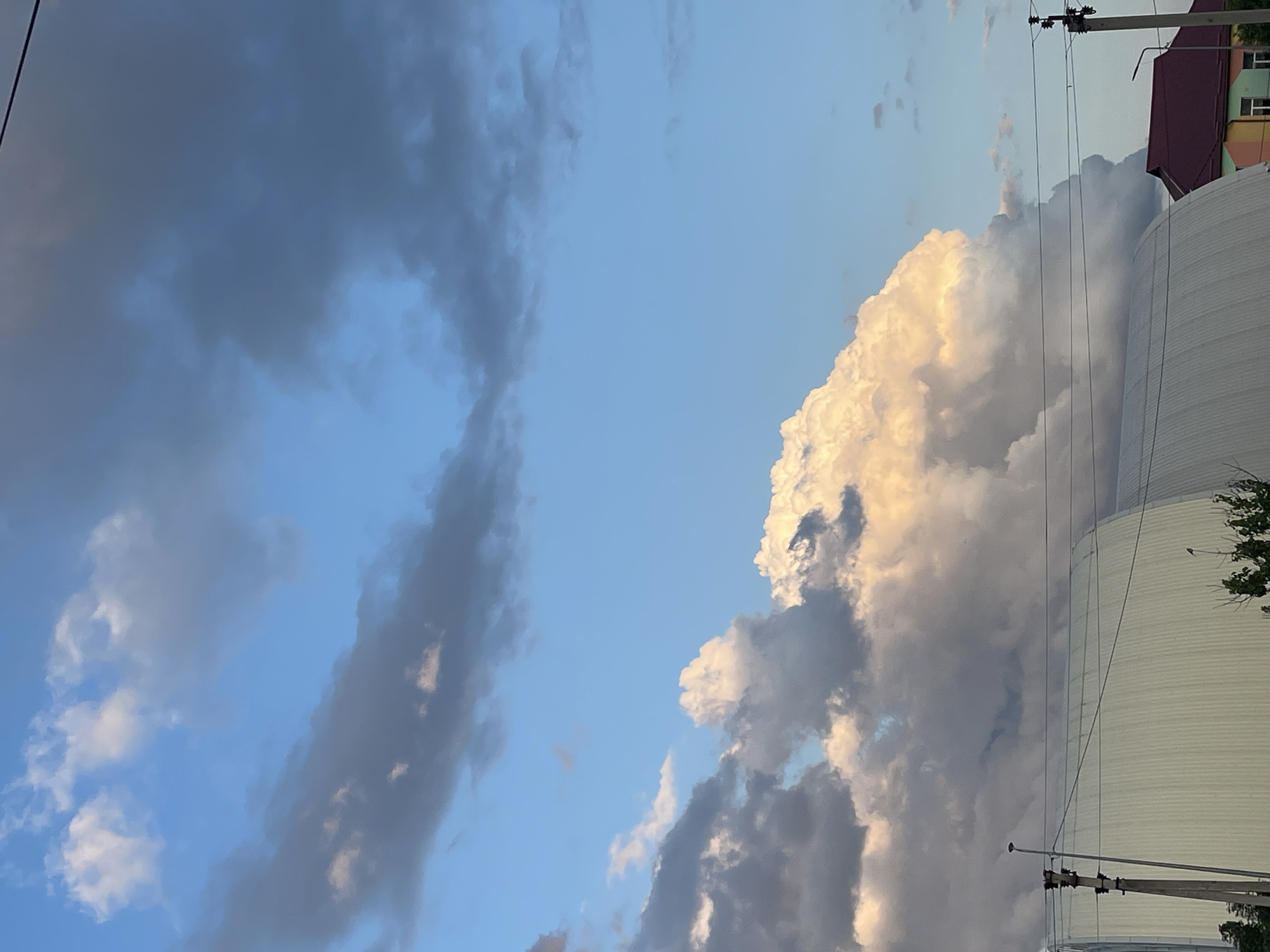 This is a vertical, eye-level, outdoor photograph taken in the city of Krasnokutsk, Ukraine. The image features a dramatic sky filled with clouds, illuminated by the warm glow of sunset. The sky is a vibrant blue, transitioning to darker, ominous gray clouds in the upper portions and to the left, suggesting an approaching storm or the remnants of one. A large, fluffy cumulus cloud dominates the center of the frame, catching the sunset light and appearing almost golden. Below the sky, a curved, light-colored building with vertical corrugated siding is visible, possibly a warehouse or an arena. A dark-colored roof of a house with a visible window is seen to the right of the large building. Overhead, power lines and utility poles stretch across the frame, indicating the presence of infrastructure. The overall scene captures a moment of natural beauty and impending weather change.
