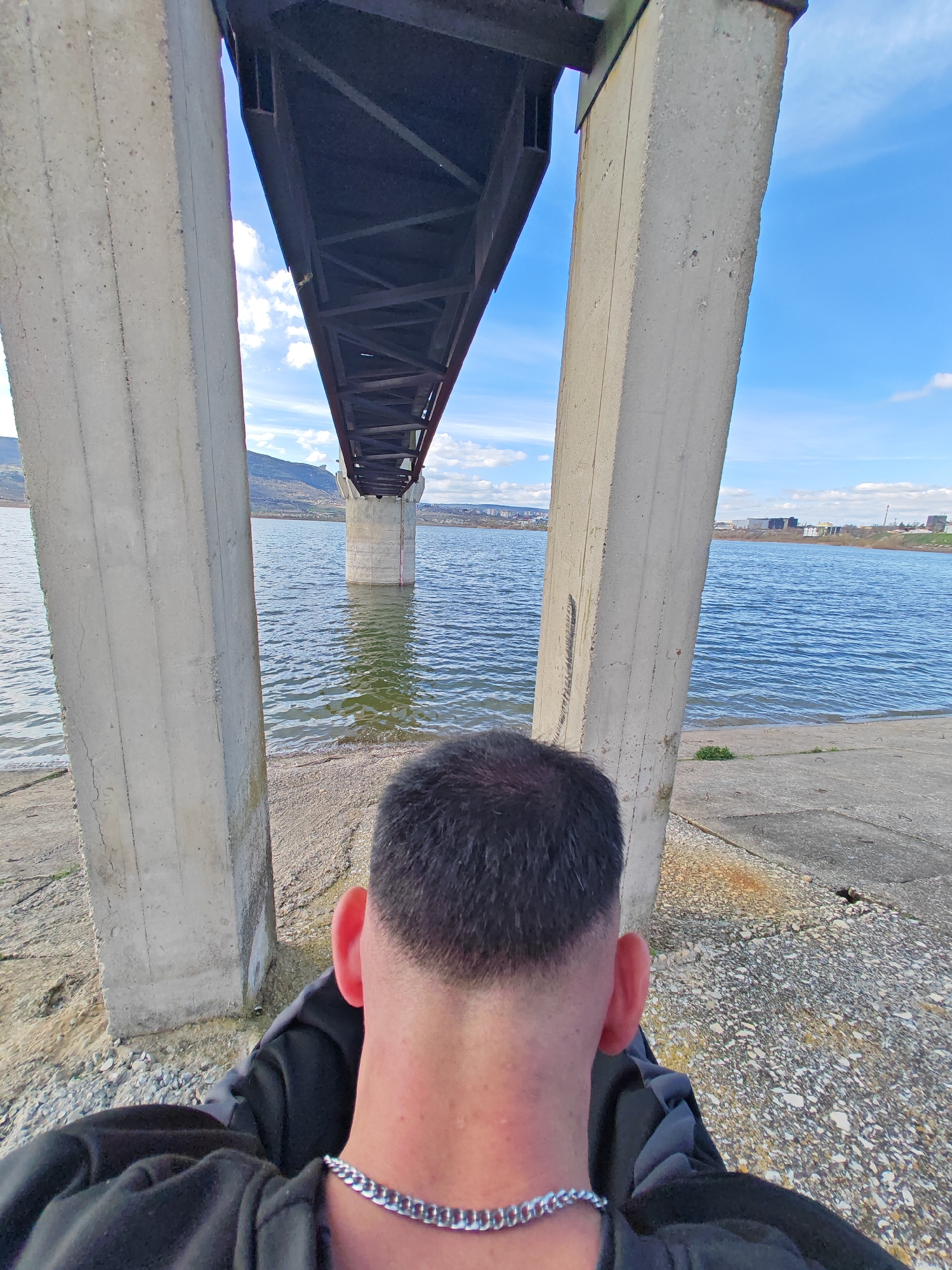 A person is seen from the back, with short, dark hair and wearing a silver chain, sitting under a bridge structure with concrete support pillars. The body of water reflects the blue sky and clouds. Beyond the water, the city of Shumen, Bulgaria is visible in the background, including a distant mountain ridge and some industrial buildings. The ground surrounding the base of the pillars consists of gravel and concrete.