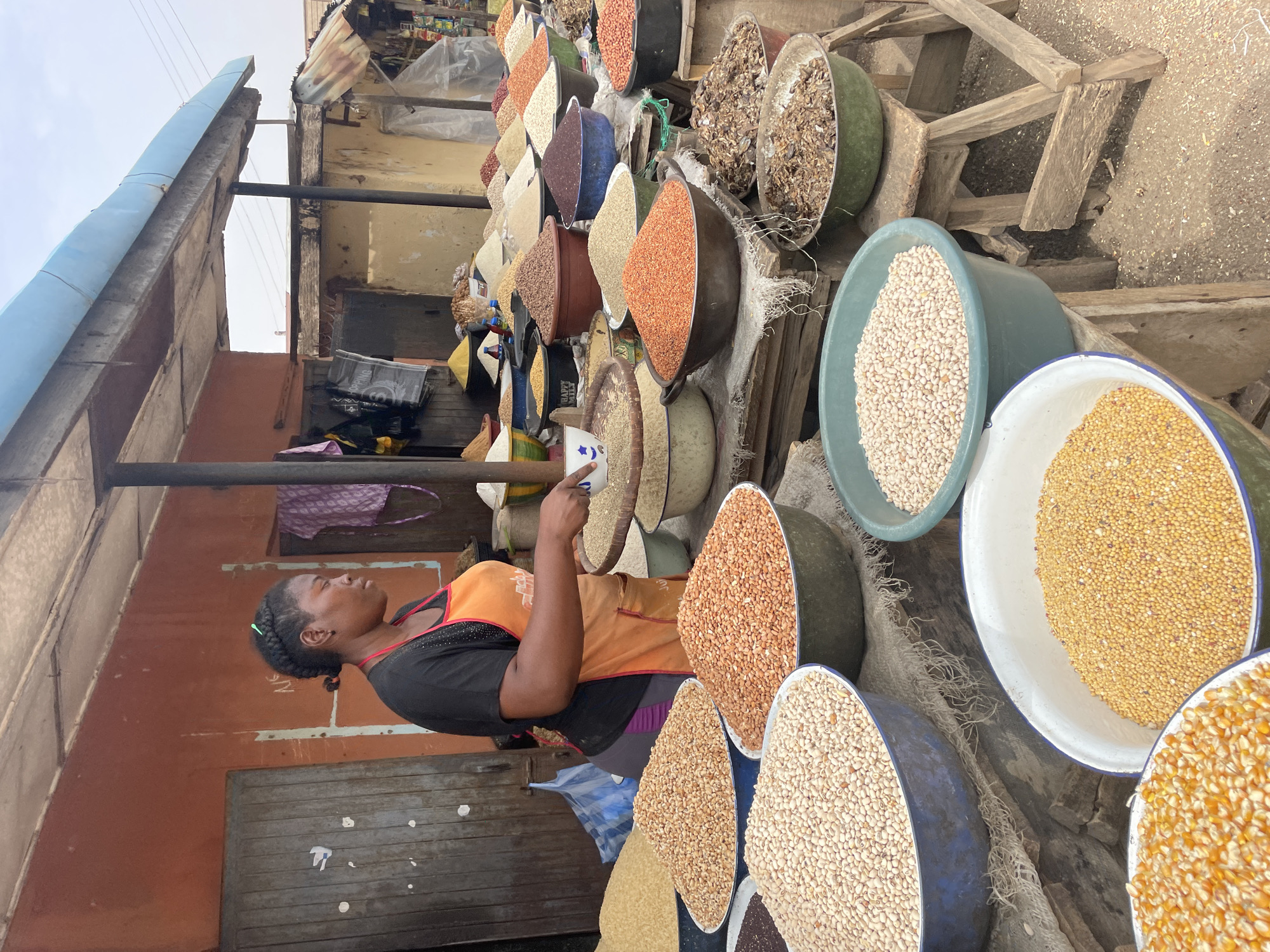 An outdoor market in Jos, Nigeria, features a woman in an orange apron serving grains or seeds to a customer. She uses a white cup to measure the product. The grains are displayed in a variety of bowls and buckets. The background includes simple structures and a partly cloudy sky.