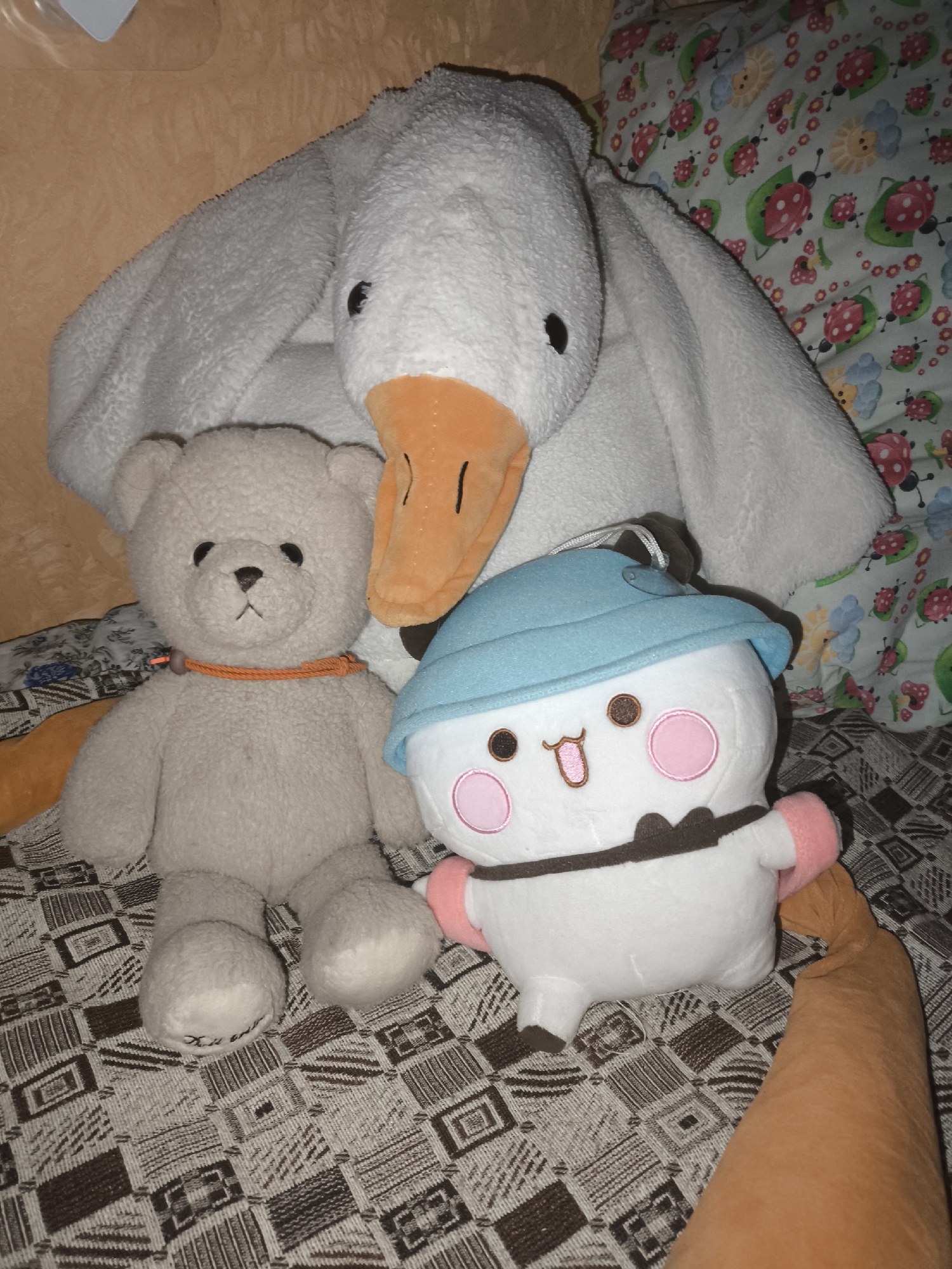 A stuffed white goose and a light brown teddy bear are positioned on a patterned blanket, possibly on a bed, in a room that appears to be in Русский Кукмор, Russia. The goose is large and its head is near the top of the frame, with an orange beak and black eyes visible. It's draped over the teddy bear, which is in the center, facing forward with a small orange string around its neck. To the right of the teddy bear is a white stuffed character wearing a blue hat and pink cheeks, with a brown collar and arms. A blurry orange stuffed limb, possibly from another toy, extends into the lower right corner of the image. The background shows a light brown textured surface and a pillow with a ladybug and flower pattern. The overall scene is dimly lit, suggesting an indoor setting at night or in low light conditions.