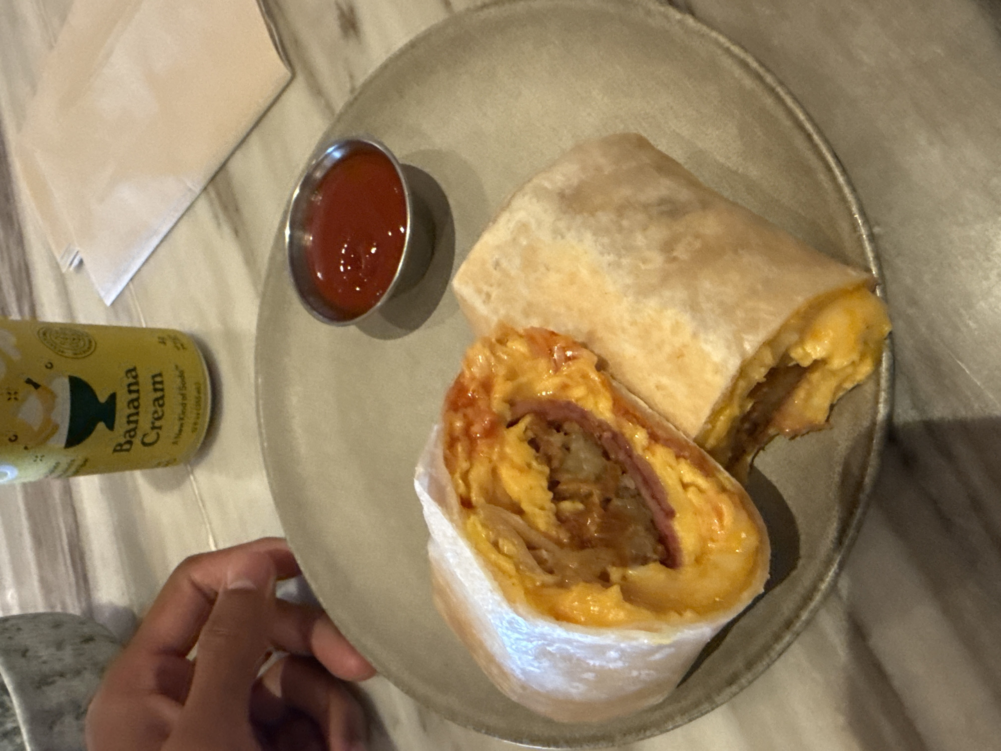 An overhead shot captures a meal consisting of two burritos, a side of sauce, and a drink can, presented on a light gray, round plate. The plate rests on a white and gray marble-patterned table. One of the burritos is partially cut open, revealing a filling of scrambled eggs, thin slices of deli meat (possibly ham or bacon), and what appears to be fried potatoes or hash browns, all encased in a light-colored tortilla. A small metal ramekin filled with a vibrant red sauce, likely ketchup or hot sauce, sits next to the burritos.
To the left of the plate, a yellow can is visible, bearing the text "Banana Cream" and an illustration of a dessert bowl. A folded white paper napkin is situated behind the plate. A person's hand, with a medium skin tone, is partially visible in the bottom left of the frame, resting near the plate. The scene suggests a casual breakfast or brunch. No specific time of day or weather is discernible beyond the context of a meal. The setting is likely indoors.