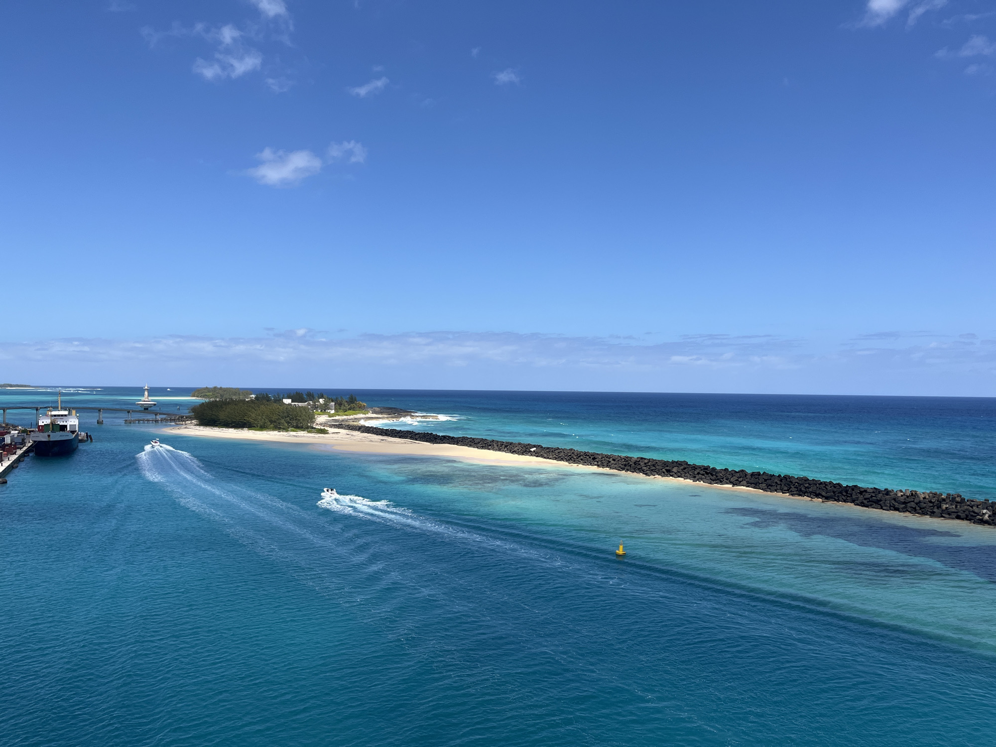 The image shows a coastal scene with a vibrant blue ocean and sky. A long, dark breakwater extends into the ocean, parallel to a sandy beach and a small island with vegetation and buildings. A channel is visible on the left side of the image, with a large cargo ship docked along a pier. A bridge crosses the channel. Two boats are moving through the water, leaving wakes behind them. A yellow buoy is located in the water near the breakwater. The location appears to be a harbor or coastal area.
