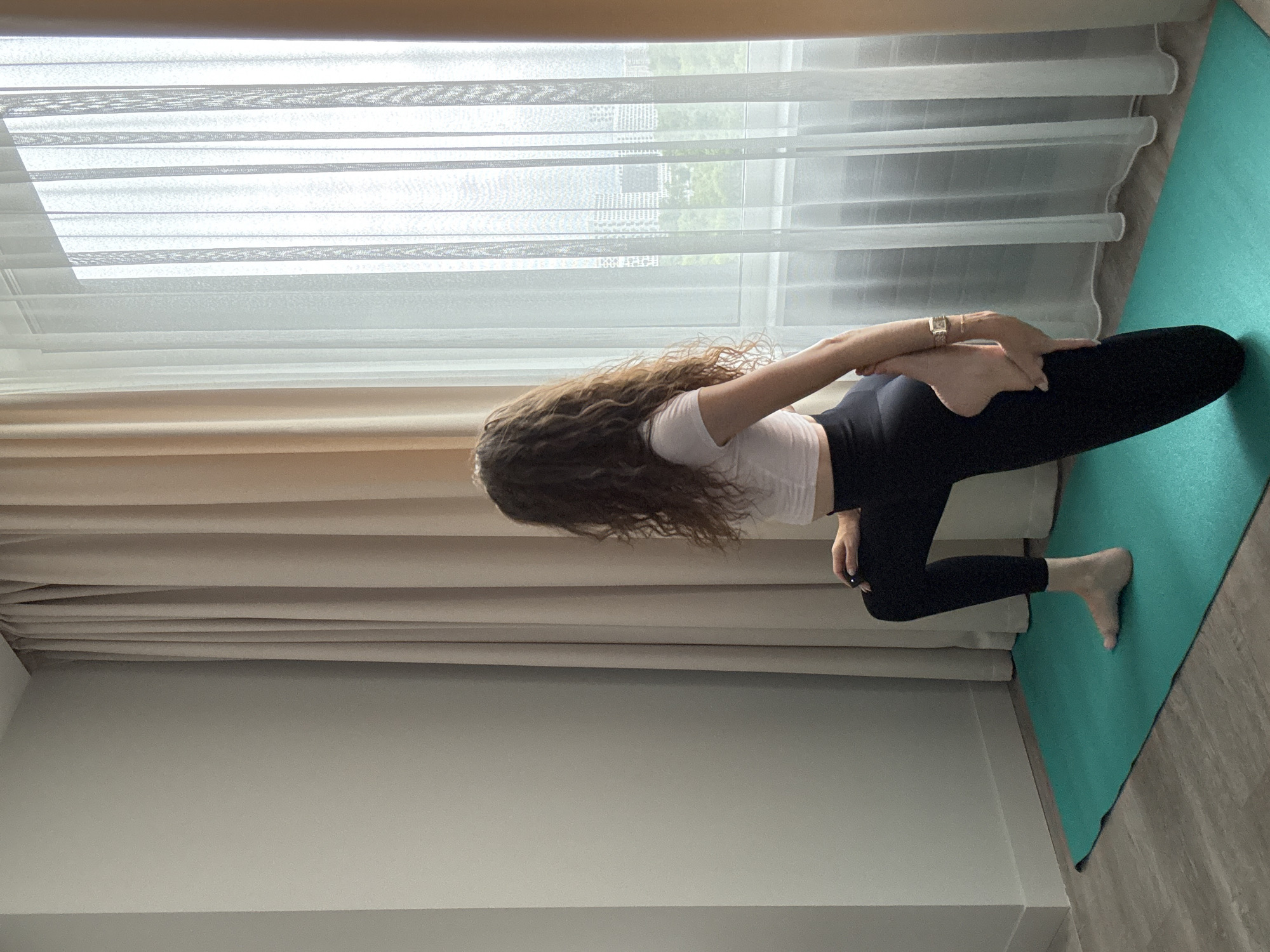 A woman with long, curly brown hair is performing a yoga pose on a teal yoga mat in a room with light-colored walls and curtains. She is wearing a white crop top and black leggings. Her right knee is bent and on the mat, while her left leg is extended behind her. She is holding her left foot with her right hand, stretching her quadricep. The room has a large window covered by sheer white curtains and thicker beige curtains. Through the sheer curtains, a cityscape with buildings and trees is visible. The floor is a light-colored wood laminate.