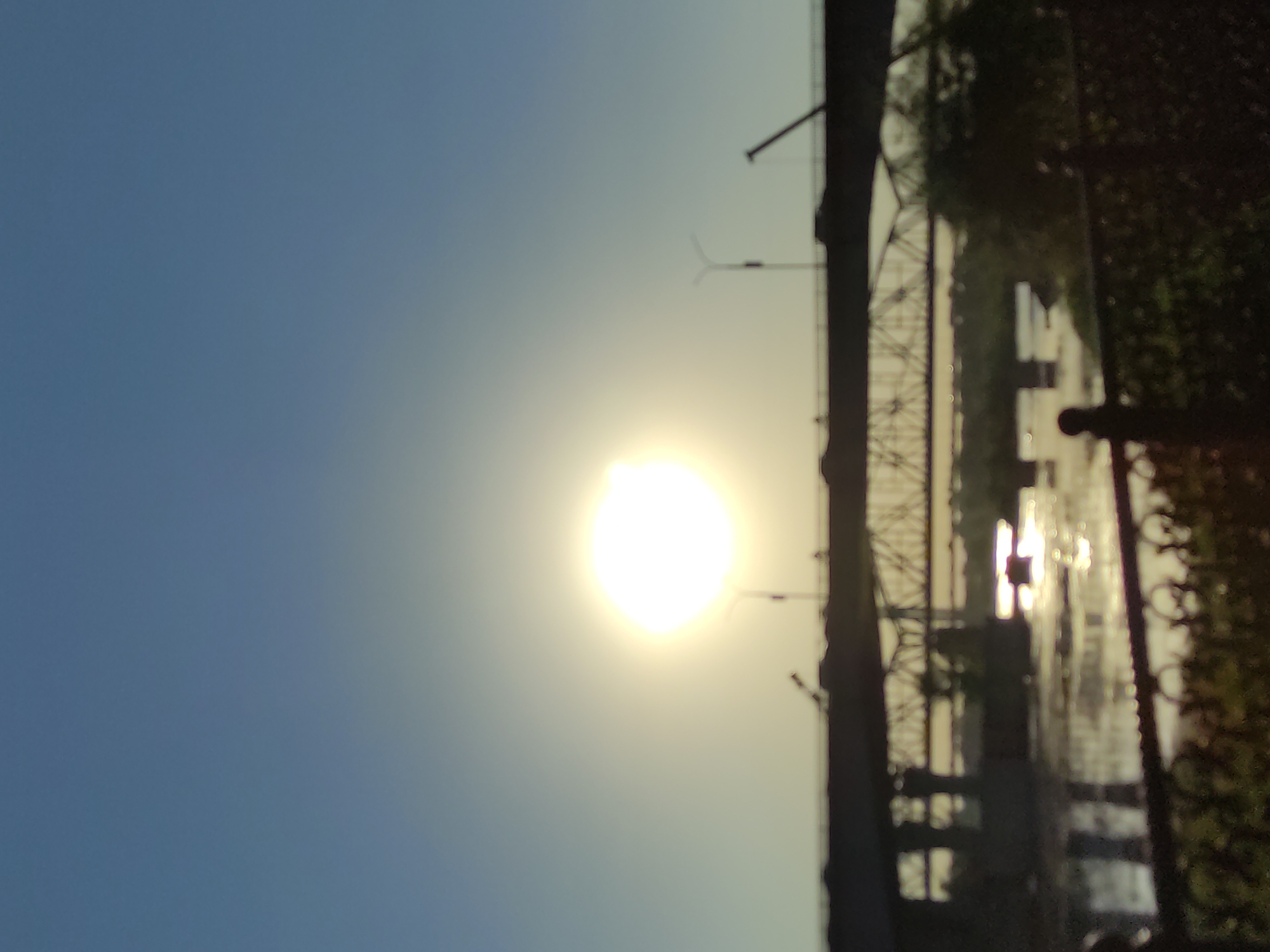 The image captures a hazy sunset over the Irtysh River in Semey, Kazakhstan. The sun, a bright, out-of-focus orb, hangs low in the clear blue sky, casting a warm glow on the scene. A prominent bridge, likely a road bridge, spans across the river, with another, older-looking truss bridge visible beneath it. Silhouettes of streetlights and possibly construction cranes are also present on the bridge. The river's surface reflects the sunlight, creating a shimmering effect. In the foreground, a dark, blurred fence with decorative metalwork and foliage suggests a public viewing area or park. The overall impression is one of a peaceful evening in an urban riverside setting.