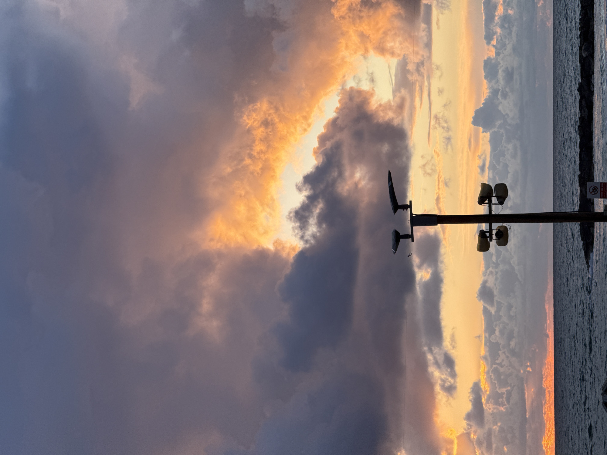 The image displays a coastal scene in Tel-Aviv, Israel, captured at sunset. The sky, occupying the upper two-thirds of the frame, features a dramatic arrangement of dense, dark grey and purple cumulus clouds. A significant portion of the right sky exhibits intense orange and yellow illumination from the setting sun, contrasting sharply with the darker cloud masses. A clear horizon line separates the sky from the Mediterranean Sea, which comprises the lower third of the image. The sea is dark blue-grey, with visible whitecaps indicating choppy conditions. A sliver of concentrated red-orange light is visible on the far left horizon, directly above the water.
In the mid-ground, a tall, dark utility pole is centrally positioned, extending from the bottom edge upwards. The pole is equipped with two dark, angular light fixtures at its top, and a small, dark bird is perched on the upper cross-arm. Below these fixtures, two sets of yellow, horn-shaped devices, possibly loudspeakers or specialized sensors, are mounted vertically on the pole. At the base of the pole, a white rectangular sign with red text, partially in Hebrew, and a red circular 'no entry' or 'no swimming' symbol is partially visible. A low, dark rock breakwater extends horizontally across the water in the lower right background. No human subjects are visible.