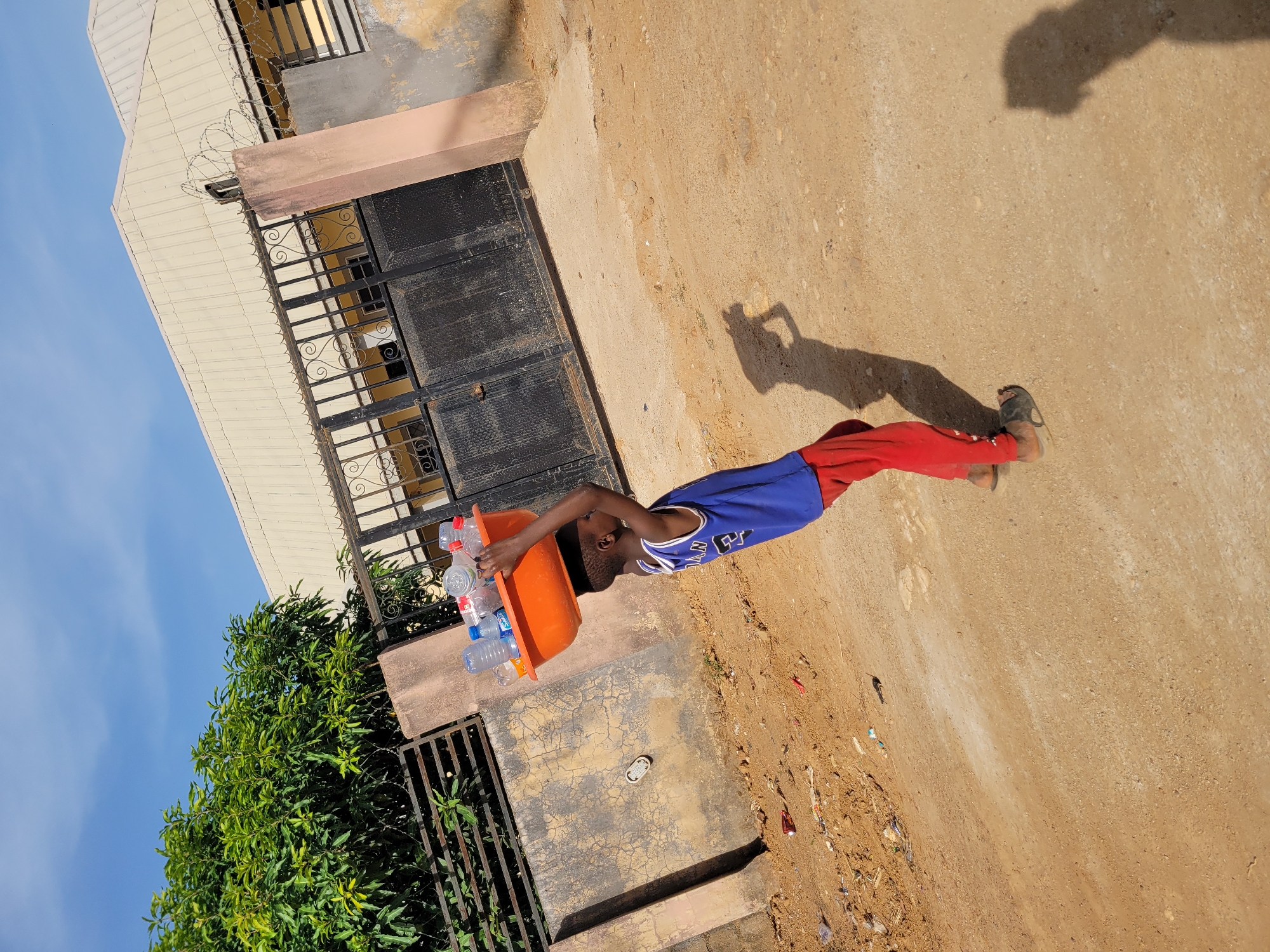 A young boy is captured outdoors, actively carrying an orange basin filled with numerous clear plastic water bottles balanced on his head. He wears a blue basketball jersey with the letters "NY" visible on the front, paired with red pants and dark sandals. His distinct shadow is cast sharply on the light brown, dusty ground, indicating bright, clear daylight, likely midday or early afternoon.
The setting appears to be a residential or semi-urban environment. In the background, a building with a light-colored corrugated metal roof, barred windows, and a dark metal gate can be seen. Barbed wire is installed along the top of a wall or fence adjacent to the building. To the left, a large, lush green tree stands against a clear blue sky. The ground is dry, uneven, and unpaved, suggesting a common street or compound area in Barakin Akawo, Nigeria. The boy's expression is neutral and focused on his task, demonstrating a common method of carrying goods in the region.