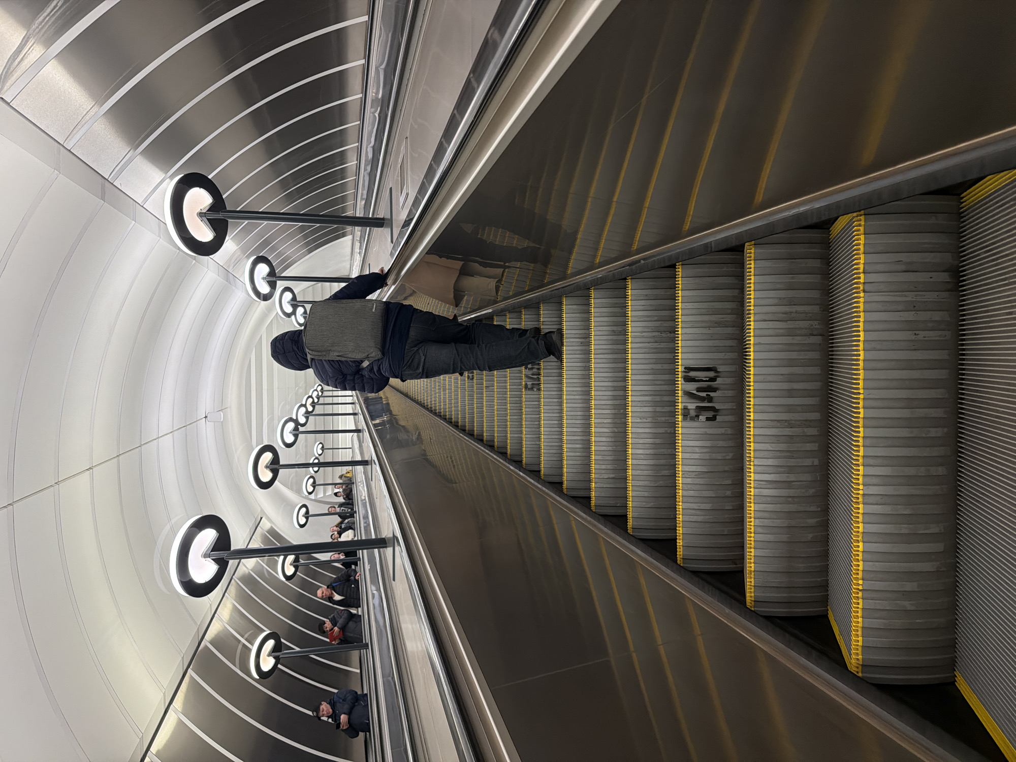 The image shows the interior of a Moscow metro station, specifically the Park Pobedy station, which is one of the deepest metro stations in the world. The scene depicts a long escalator descending into the station. A person wearing a dark jacket and carrying a backpack is riding the escalator. Other people can be seen behind this person, also riding the escalator. The walls of the station are characterized by a unique design featuring white panels with gray stripes and circular lighting fixtures resembling Pac-Man figures. The escalator steps are gray with yellow edges. The number "540" is visible on one of the steps.