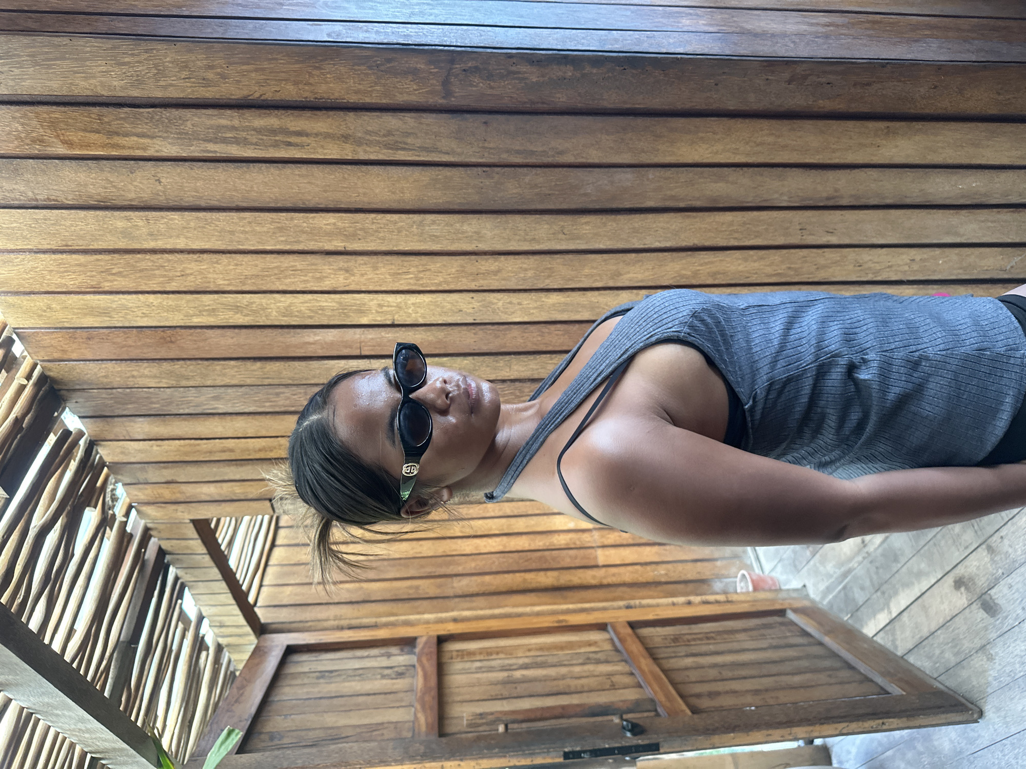 A woman is standing and posing for a photo. She is wearing a grey ribbed tank top and black sunglasses. She has her hair pulled up in a messy bun. She is standing in front of a wooden wall, which is part of an outdoor structure. The structure appears to be made of natural materials, with bamboo poles forming a roof. The floor is made of wooden planks. There is an open wooden door to the left of the woman, and a red bucket can be seen through the doorway. The scene is likely in a tropical outdoor setting, possibly a resort or a cafe in North Kuta, Indonesia.