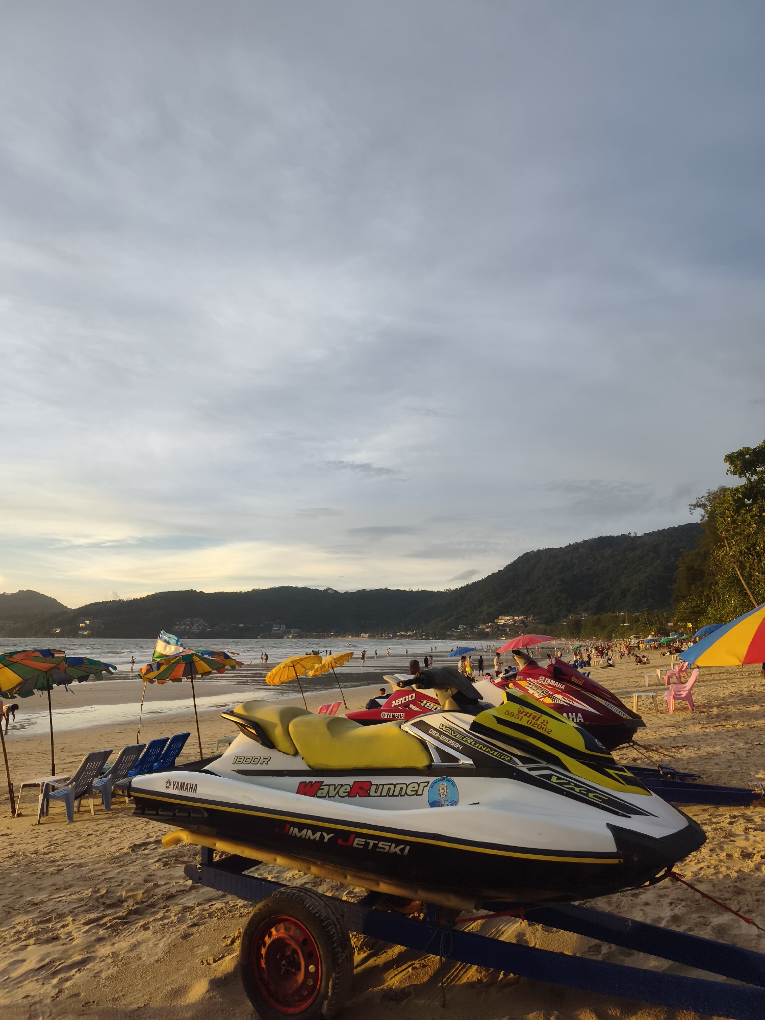 The image depicts a scene on Patong Beach in Thailand. In the foreground, two jet skis are parked on a sandy beach, resting on trailers. One jet ski is primarily white with yellow accents and the "WaveRunner" logo. The other, partially visible behind it, is red. Several beach chairs and umbrellas, in various colors like yellow, orange, green, and blue, are scattered on the sand.
In the middle ground, the beach is populated with numerous people walking, sitting, and engaging in various activities. Beach umbrellas dot the area, providing shade. The sea is visible, with gentle waves rolling towards the shore.
The background features a hilly landscape covered in lush green vegetation, with buildings visible on the slopes. The sky is overcast with soft clouds, and the lighting suggests it is either early morning or late afternoon, with the sun casting a warm glow. The overall atmosphere is that of a typical beach resort scene.