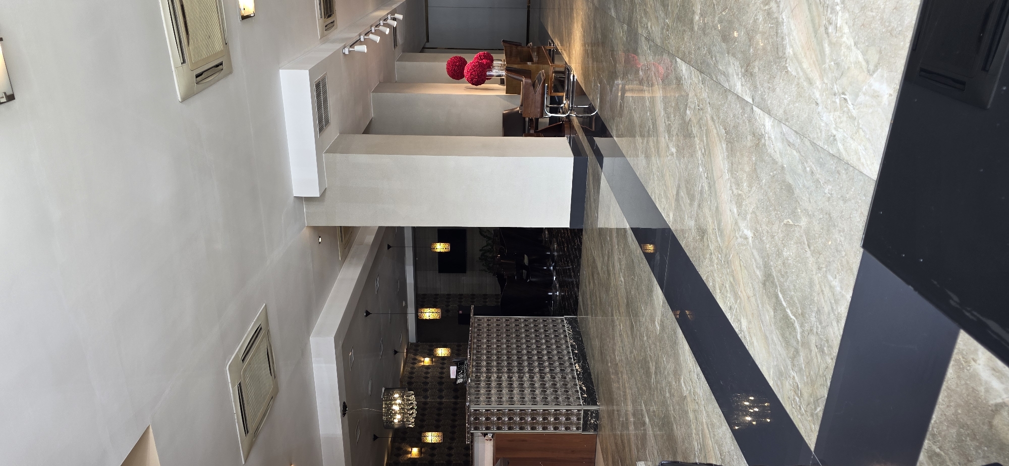 This is a photograph of a hotel lobby in Bishkek, Kyrgyzstan. The lobby features a polished marble floor with a dark inlaid stripe. To the left, a reception desk is visible, with a decorative metal front. Above the desk, a crystal chandelier hangs, surrounded by a series of pendant lights and sconces. In the background, there are dark upholstered chairs and tables, suggesting a seating area. To the right of the frame, a large beige pillar stands, supporting the ceiling. Nearby, a collection of modern chairs are arranged, and a vase holding bright red floral arrangements is placed on a surface. The ceiling is white and features several air conditioning vents and recessed lighting fixtures. The overall impression is one of modern, clean, and somewhat luxurious interior design.