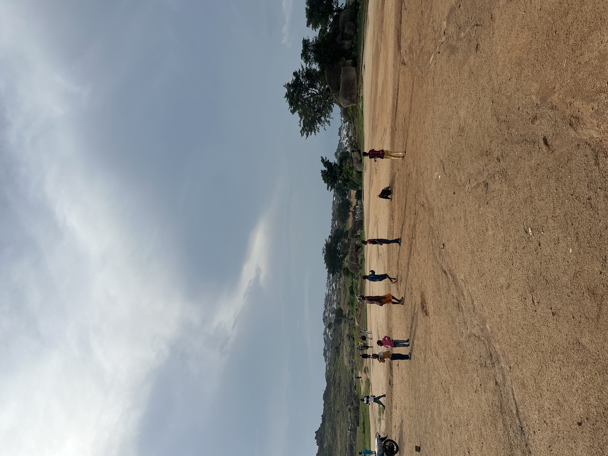 The image depicts an outdoor scene in a somewhat arid, hilly environment. The foreground is a wide expanse of dry, reddish-brown dirt or sand, with some scattered rocks and tire tracks visible. In the middle ground, a group of people, predominantly children, are scattered across the open area. Several children are standing or walking, and some appear to be engaged in activity, though specific interactions are not clearly discernible. One child is seen crawling on the ground.
Further in the background, there is a cluster of trees and large rocks, characteristic of the rocky outcrops found in Jos, Nigeria. Beyond the trees and rocks, a settlement with buildings featuring white roofs is visible, suggesting a nearby town or village. The sky is overcast with dramatic, grey clouds, indicating a potentially stormy or changing weather condition. A motorcycle is parked on the left side of the foreground. The overall impression is of a rural or semi-urban landscape in Jos, Nigeria, during daylight hours under a cloudy sky.