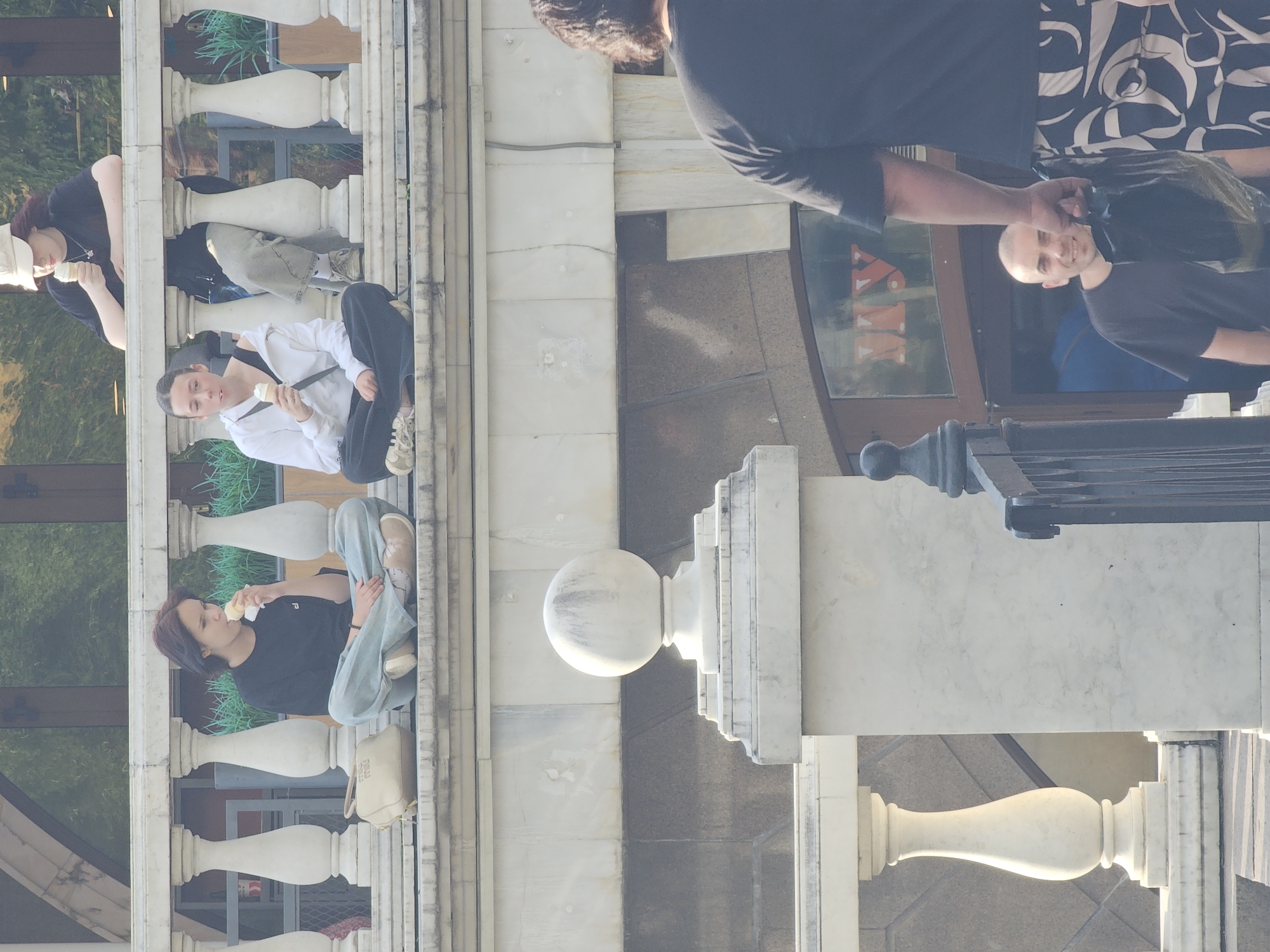 The image shows an outdoor scene in Moscow, Russia, featuring several individuals. In the foreground, a person with short dark hair and a black t-shirt is partially visible from the back, holding a black plastic bag. To their left, a man with a shaved head and a dark t-shirt is smiling and looking towards the camera. Behind them, a glass-paneled entrance with the letters "MY" visible is part of a building.
In the middle ground, two young women are sitting on a stone ledge. The woman on the left has dark hair and is wearing a black t-shirt and light blue pants, holding an ice cream cone. The woman on the right has dark hair pulled back and is wearing a white hoodie over a black top and dark pants, also holding an ice cream cone. Next to the woman on the left is a light-colored handbag.
In the background, another person wearing a light-colored baseball cap and a dark t-shirt is visible, also holding an ice cream cone. They are sitting on a stone balustrade, with lush green foliage visible behind them. The architecture suggests a public space with decorative stone elements.