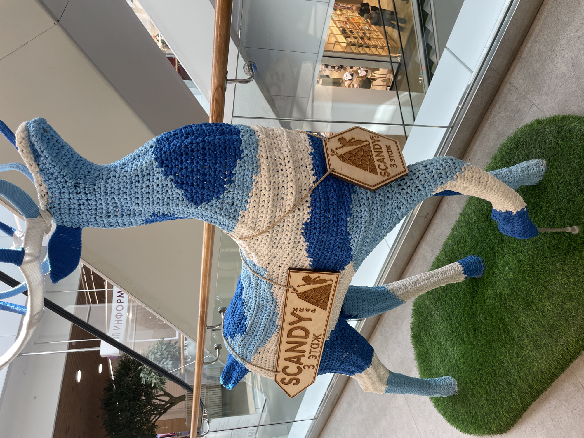 Here is a detailed description of the image:
The image shows a large, crocheted sculpture of a reindeer in a shopping mall. The reindeer is primarily blue and white, with horizontal stripes. It has a wooden sign hanging from it, with the words "Scandy Park 3 этаж" (Scandy Park 3rd floor) on it. The reindeer is standing on a patch of artificial grass. The mall has glass railings and multiple floors. There is a shop visible in the background. The photo was taken in the city of Izhevsk, Russia, as indicated by the Cyrillic script on the sign.