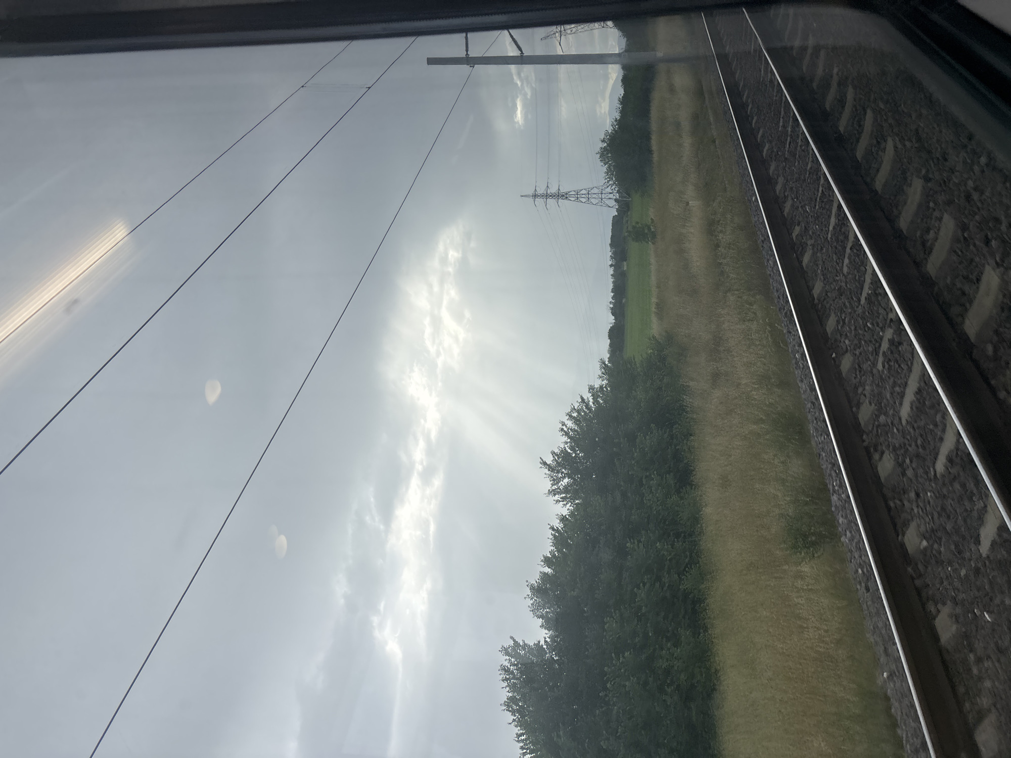 A landscape scene captured from the window of a moving train, oriented vertically. On the right side of the frame, two parallel train tracks with ballast and sleepers recede into the distance. To the left of the tracks, a strip of brownish-green grass and an expansive field extend, bordered by a dense line of green trees. Above the trees and field, the sky is partly cloudy, with striking crepuscular rays (sunbeams) piercing through the clouds, illuminating parts of the landscape. Several power lines stretch across the mid-ground, supported by two prominent high-voltage pylons visible against the sky and trees. The image's left side shows reflections, indicating it was taken through a window. The overall impression is one of travel through a rural or semi-rural area. The time of day appears to be daytime, possibly morning or late afternoon given the angle of the sun's rays. No specific human activity or people are visible. No text is discernible in the image. The location context provided is Sinitovo, Bulgaria.