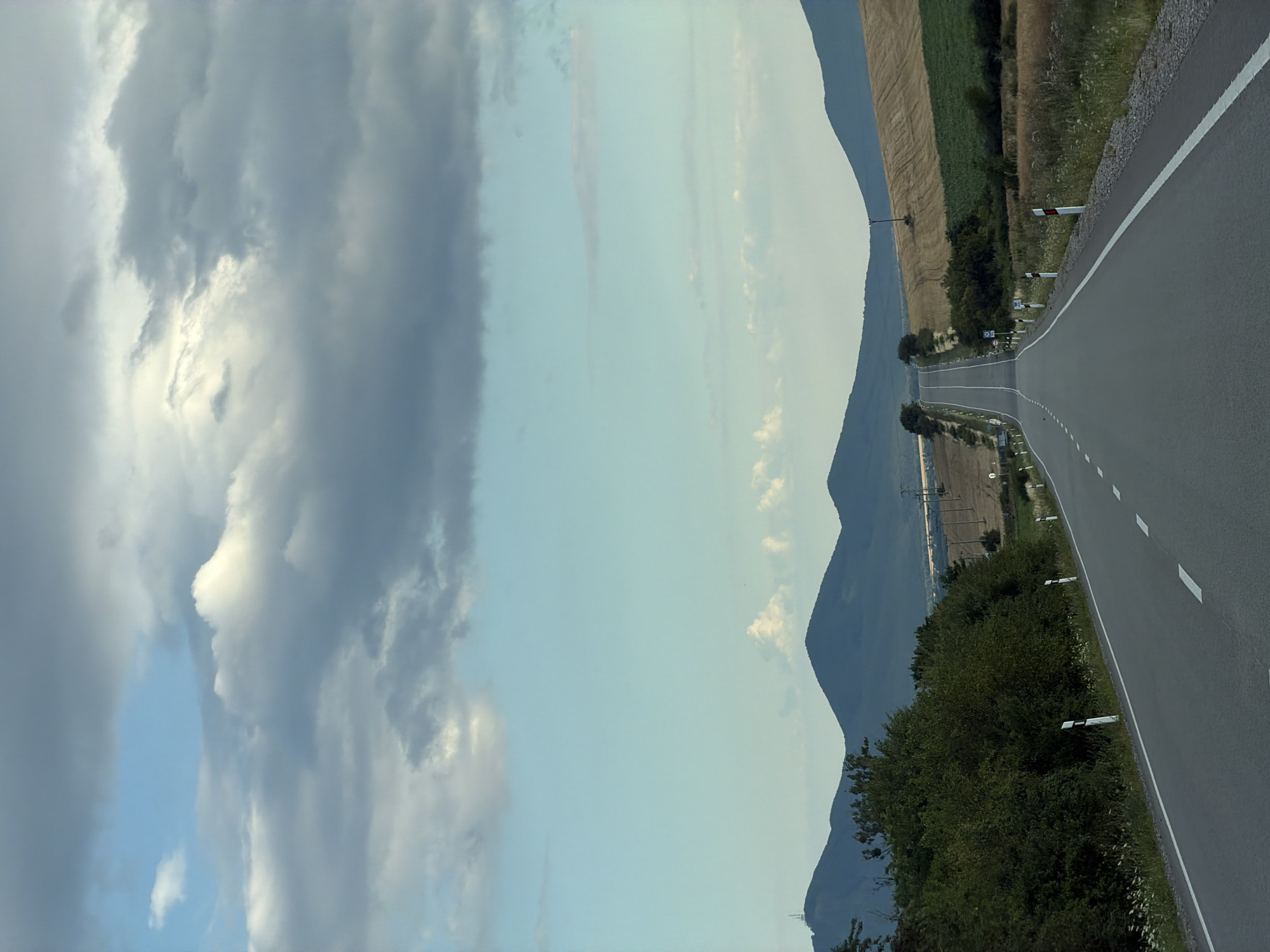 The image depicts an empty asphalt road stretching into the distance towards rolling hills and mountains under a cloudy sky. The road is lined with white and red reflective markers. The surrounding landscape consists of fields, vegetation, and scattered trees. In the background, a prominent peak with what appears to be a communication tower is visible. The scene is devoid of any people or vehicles, suggesting a quiet, rural setting. The weather appears overcast but not actively raining. This location is identified as Luhyňa, Slovakia.