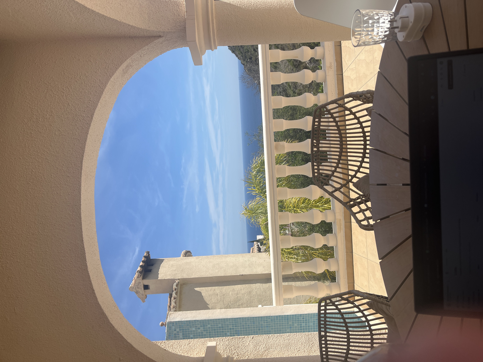 The image shows a view from a balcony in el Poble Nou de Benitatxell / Benitachell, Spain. A stone arch frames the blue sky and a view of the Mediterranean Sea. A white balustrade runs along the balcony, separating it from the view. Two wicker chairs sit around a wooden table. A tablet and a glass with an electrical outlet attached sit on the table. In the background, part of a nearby building is visible, featuring a chimney, tiled accents, and surrounding vegetation.