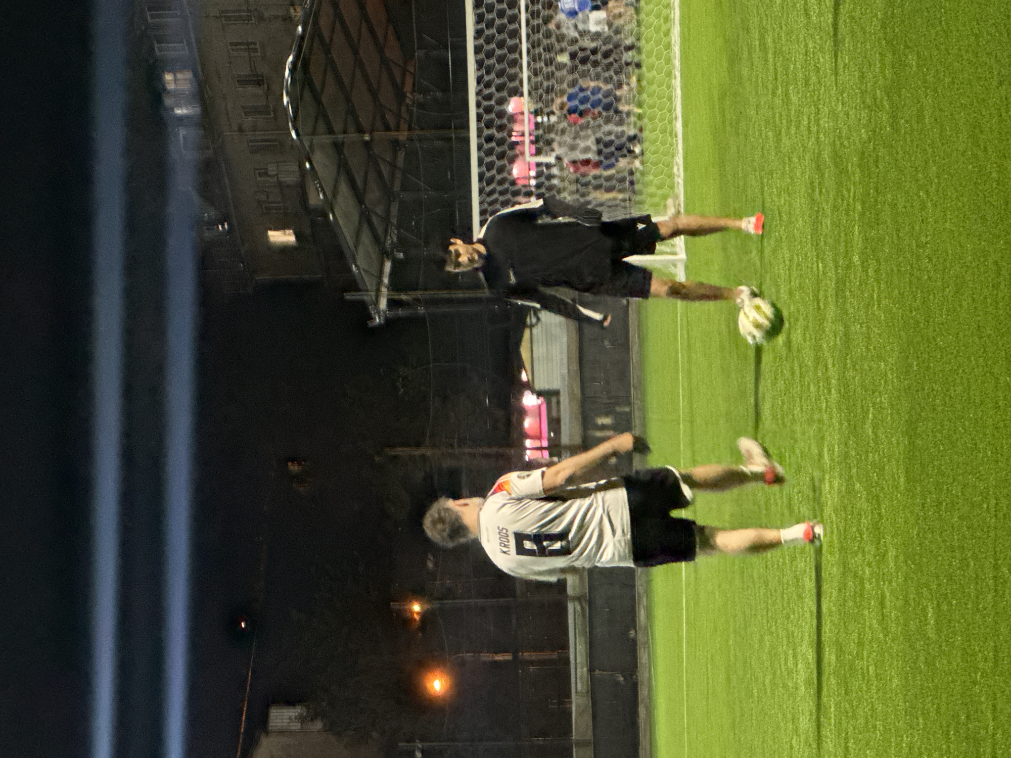 An outdoor soccer game or practice is captured at night on a vibrant green artificial turf field. Two male players are visible under artificial lighting. In the foreground, a player is seen from behind, wearing a white jersey with the name "K. ROOS" printed above the number "9", paired with black shorts. Further back, closer to a white soccer goal with a black net, another player stands in a black long-sleeved jersey and black shorts, with a soccer ball near his feet. The background reveals an urban setting with dark buildings, some displaying illuminated windows. Bright, artificial light beams streak across the left side of the image, indicating powerful stadium lights illuminating the field. The scene depicts an active sporting event taking place after dark in what is contextualized as Tbilisi, Georgia.