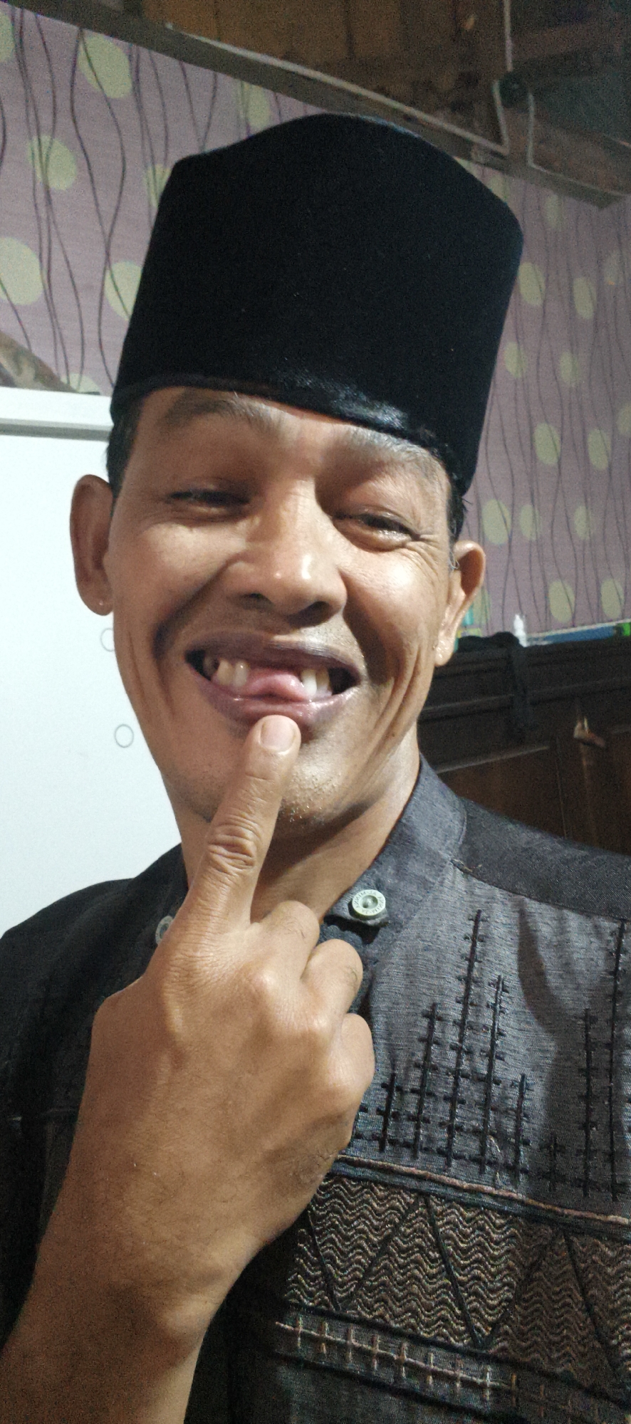 The image shows a close-up, head-and-shoulders portrait of a man wearing a black peci (a traditional cap worn in Indonesia) and a dark, patterned shirt. He is looking at the camera with his mouth open, tongue visible, and his index finger touching his lip. The background appears to be an indoor setting with patterned wallpaper and some shelving. Based on the man's attire and facial features, the image is likely taken in Muaro Jambi, Indonesia.