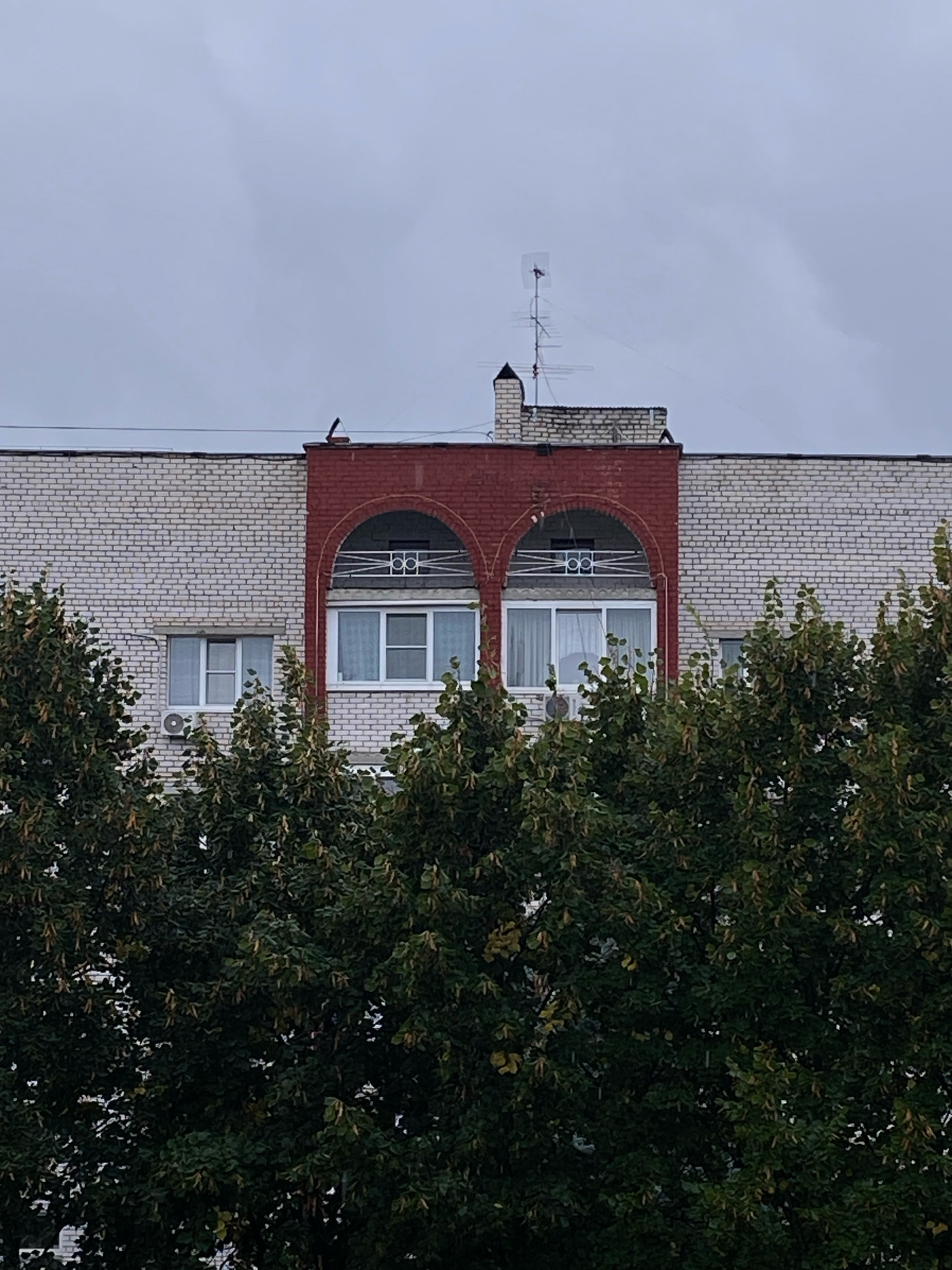 The image captures the upper facade of a multi-story apartment building in Ivanteyevka, Russia, on a cloudy day. The building is constructed with light-colored brick, with a central section clad in dark red brick. This red section features two arched openings, each containing a balcony with decorative white railings above a set of windows. To the left and right of the red section, the building is also faced with light brick. On the red brick section, there is a small chimney with a decorative cap and a television antenna. Dense green foliage from trees obscures the lower portion of the building.