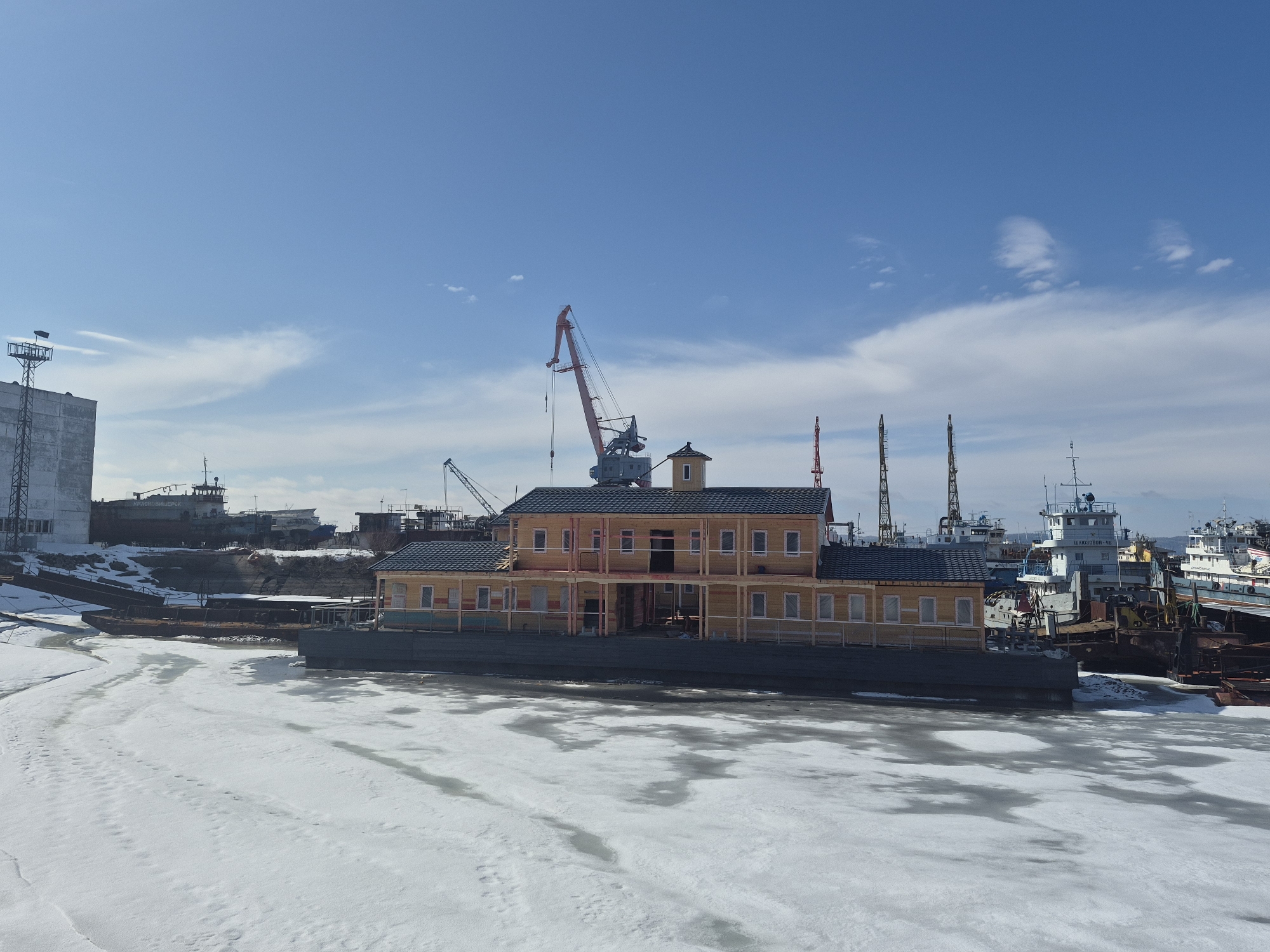 The scene depicts a partially frozen body of water with a yellow two-story building floating on a barge in the foreground. The building has a dark roof, numerous windows, and visible internal framing. A construction crane is visible behind the building. In the background, there are various ships and industrial structures, including a tall building and other cranes. The sky is light blue with scattered clouds. The location appears to be an industrial waterfront area, likely a shipyard.