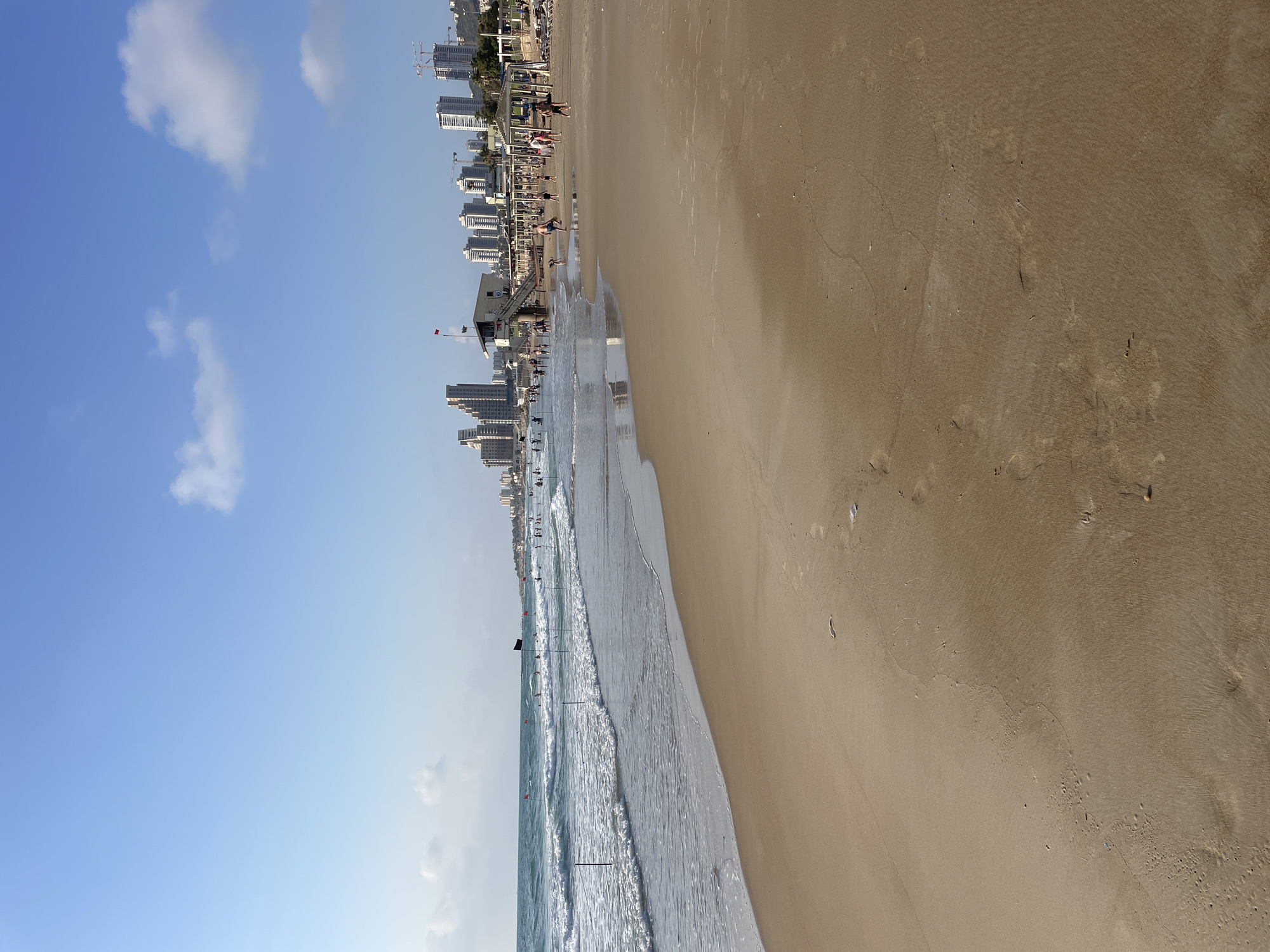 The image depicts a sunny day at a beach in Haifa, Israel, with the Mediterranean Sea on the left and a sandy beach in the foreground and middle ground. The water exhibits gentle waves and several people are visible swimming or wading. Numerous small, red flags on poles are planted in the water, likely marking swimming zones or hazards. A black flag is also visible.
In the middle ground, a lifeguard station with a red flag on top is situated near the shore. A cluster of people are gathered around the station and on the beach in front of it, with some walking and others standing. Further back, a row of buildings, including modern high-rise apartments and hotels, lines the coast, indicating an urban setting. Construction cranes are visible among some of the buildings, suggesting ongoing development. The sky is clear blue with a few scattered white clouds. The sand on the beach shows footprints and some debris.