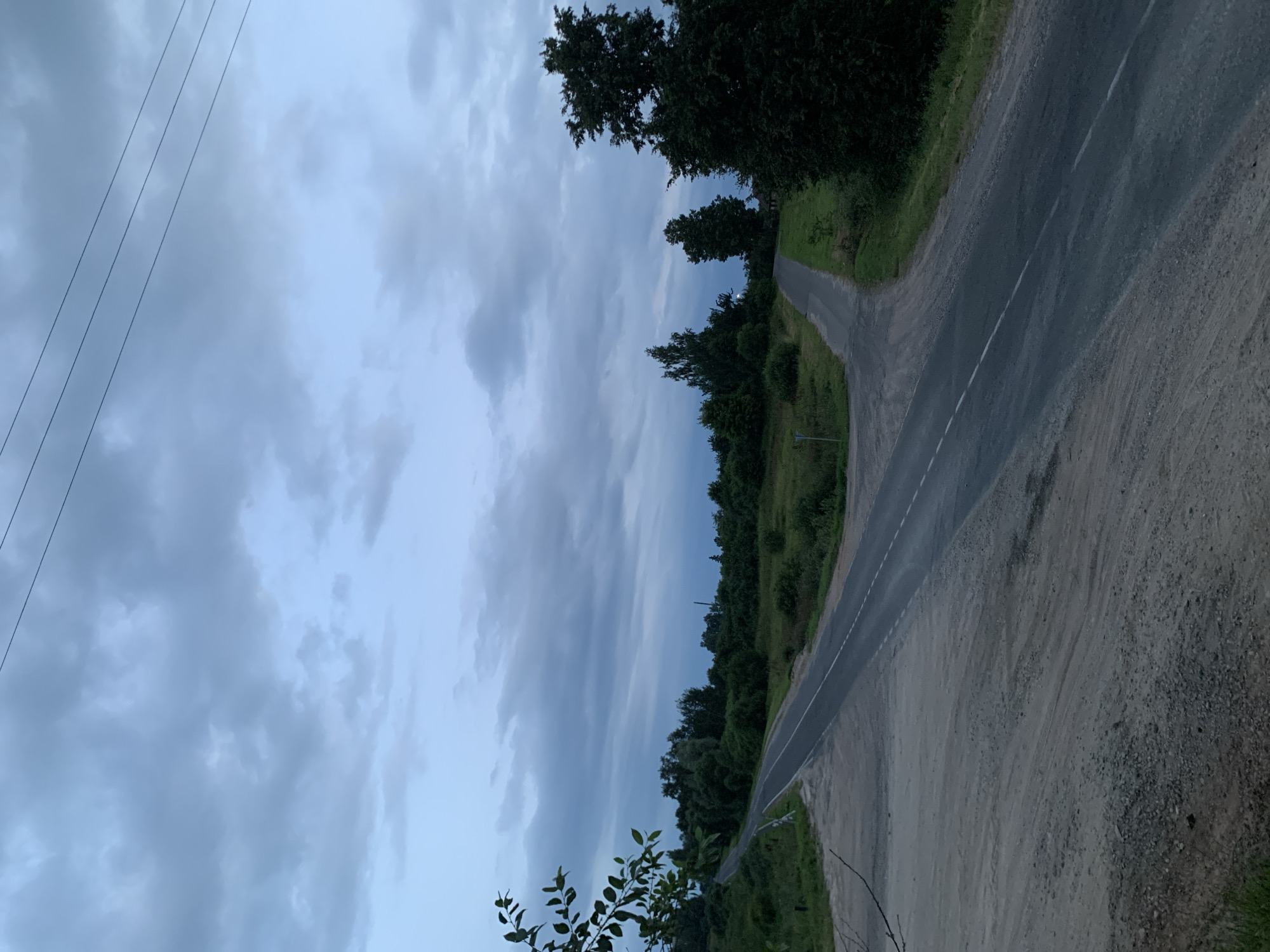 The image depicts a rural road intersection under a cloudy sky in Відзаўскі сельскі Савет, Belarus. The road is asphalt with a white dashed line down the center, splitting into two paths. The left path leads uphill and curves to the left, bordered by green grass and trees. The right path curves to the right, also surrounded by vegetation. The foreground is a dirt and gravel area. Power lines are visible against the sky. The overall atmosphere is overcast and natural, with no people or vehicles present.