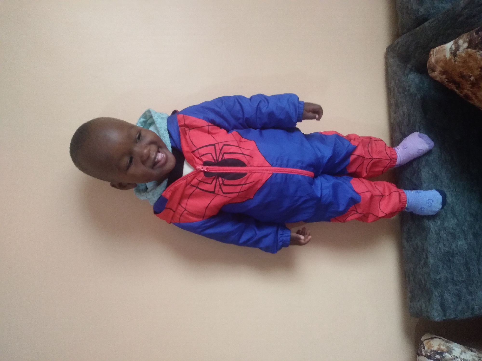 A young child wearing a blue and red Spiderman costume, complete with a hood, stands on a dark green couch in front of an orange wall. The child is smiling and looking down. The child's left foot is visible, wearing a light blue sock. The right side of the image shows part of another couch cushion. The location is a domestic indoor setting in Nyeri, Kenya.