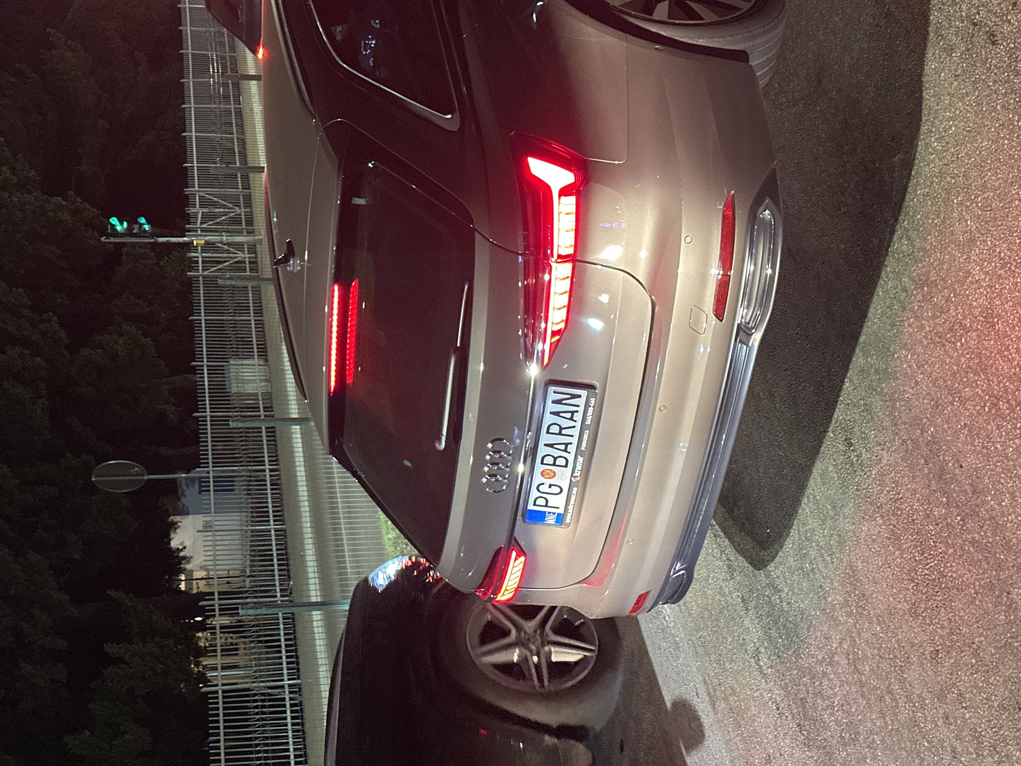 The image shows the rear of a beige Audi A6 Avant parked at night. The license plate reads "PG" followed by "BARAN". The car is in a city street, likely in Budva, Montenegro, given the license plate format. In the background, there is a white fence and trees. A black car is partially visible on the left. A traffic light with green signals is visible in the distance. The scene appears to be a typical urban street at night.