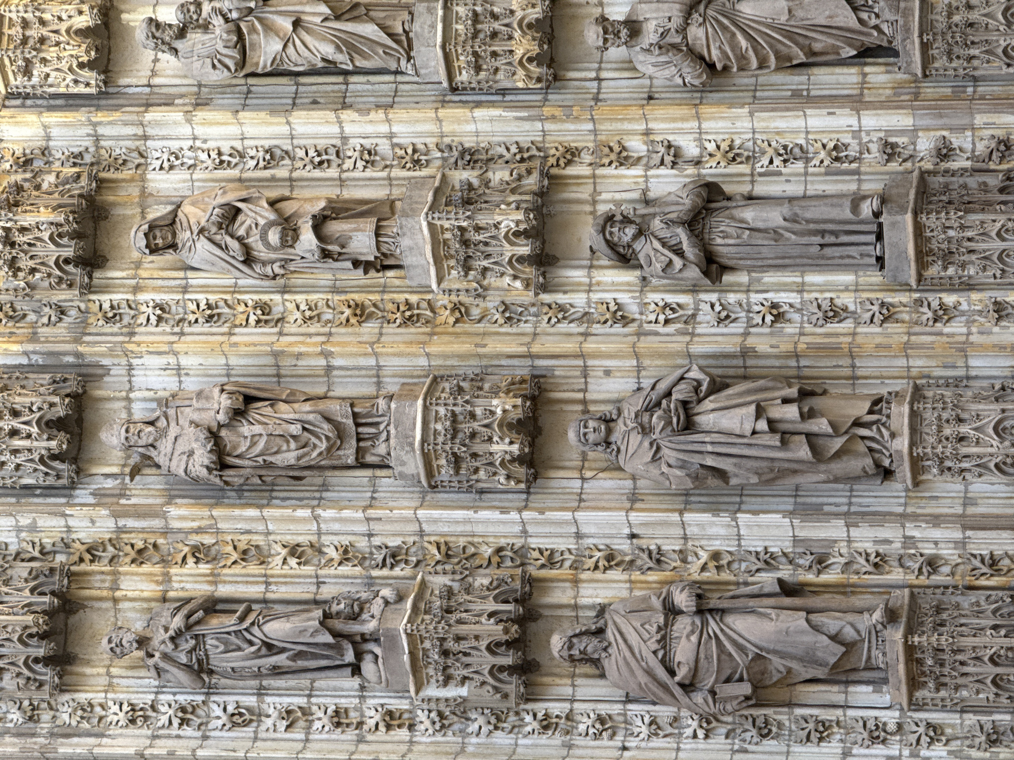 This media file showcases a section of the exterior facade of a grand Gothic building, likely a cathedral, in Seville, Spain. The image focuses on several sculpted effigies of people, arranged in horizontal rows against a backdrop of intricate stone carvings. These effigies are recumbent figures, appearing to be historical or religious figures, possibly saints or royalty.
In the top row, a sculpted figure lies on its back, with its head resting on a pillow and hands clasped over its chest. Another figure is similarly posed to its right. The second row from the top features a figure lying on its side, its head turned towards the viewer, holding a small orb or child in its arms. To its right, another figure is depicted in a similar recumbent pose. The third row continues this pattern with two more sculpted figures lying horizontally. The bottom row shows two more figures, one with its head slightly raised and the other in a relaxed reclining posture.
The figures are carved in stone and appear weathered, suggesting they are ancient. They are dressed in long, draped robes, characteristic of medieval or Renaissance attire. The architectural elements surrounding the figures are equally ornate, featuring detailed stonework, ornamental borders with floral motifs, and Gothic tracery. The lighting suggests it is daytime, with natural light illuminating the stone surfaces. There are no visible text elements in the image. The overall impression is one of solemnity and historical grandeur, typical of religious or monumental architecture found in historic European cities.