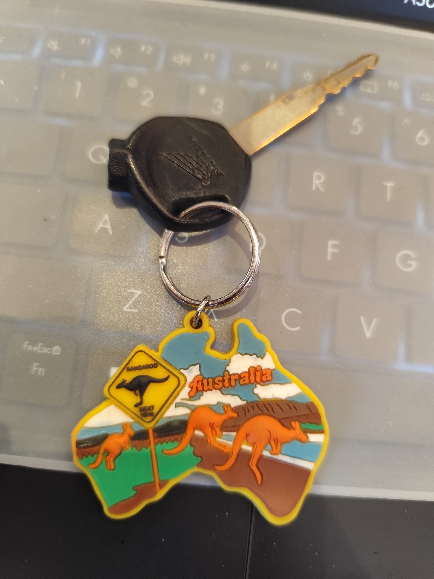 A keychain shaped like the continent of Australia with kangaroos and the word "Australia" is hanging from a car key. The keychain and key are placed on a grey keyboard.