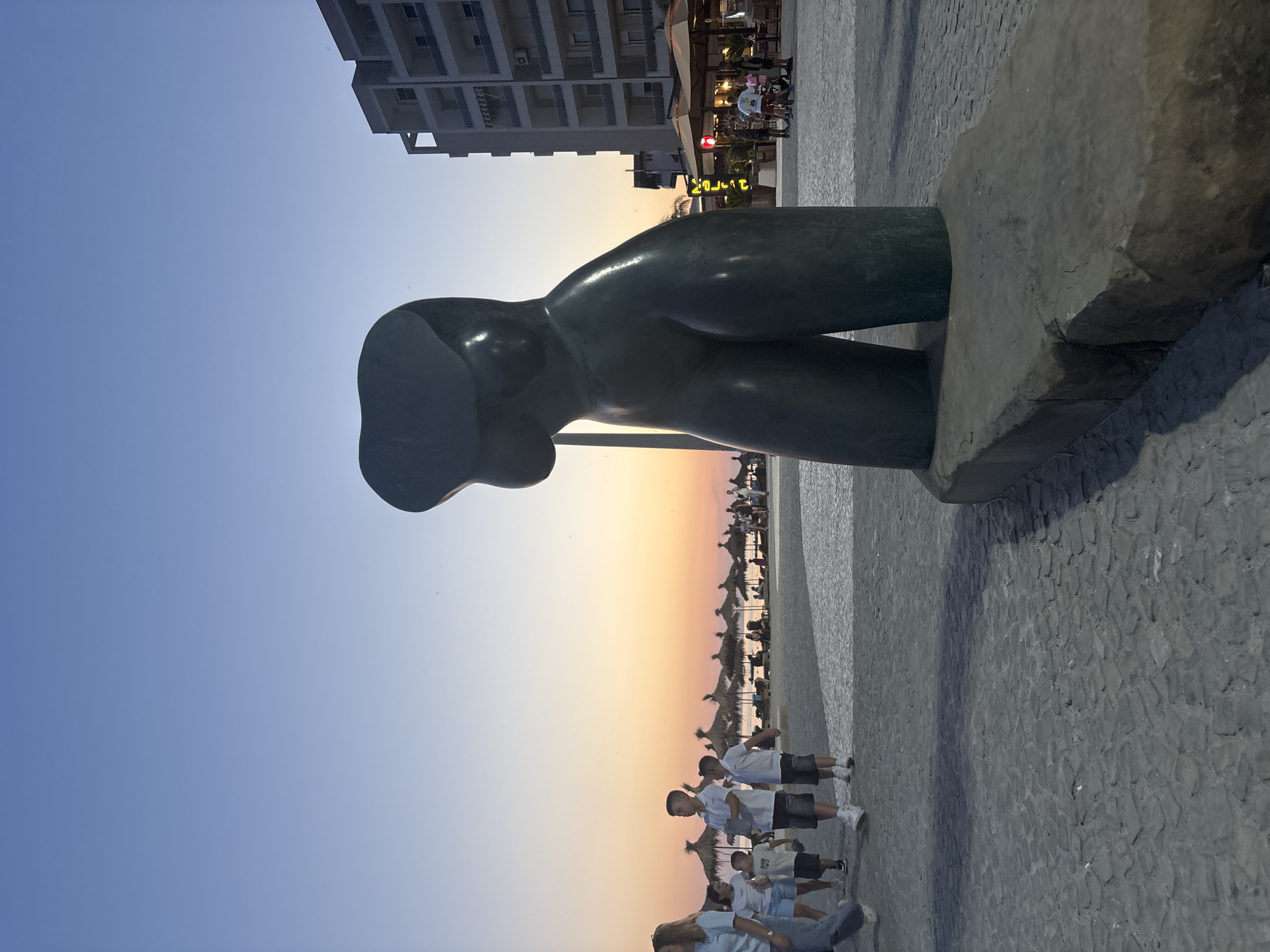 The image captures a scenic view of a beach promenade in Bashkia Durrës, Albania, during sunset. In the foreground, a large, dark green, abstract sculpture of a human torso stands prominently on a stone base. The sculpture is centrally positioned and its curved form dominates the lower right portion of the frame.
The background reveals a bustling beach scene. A paved walkway, made of grey cobblestones, stretches across the foreground and leads towards the beach. Several people, including adults and children, are walking along the promenade. Some are dressed in casual summer attire.
Beyond the promenade, rows of thatched sun umbrellas are visible on the sandy beach, indicating a popular leisure spot. The sea is a calm expanse, merging with the horizon. The sky is painted with the warm hues of sunset, transitioning from a soft orange and pink near the horizon to a clear blue at the top of the frame.
To the right of the sculpture, a multi-story building with balconies and windows stands tall, suggesting an urban backdrop to the beach setting. In front of the building, there are outdoor seating areas with umbrellas, hinting at cafes or restaurants. The overall atmosphere is peaceful and idyllic, capturing the end of a pleasant day at the seaside.
