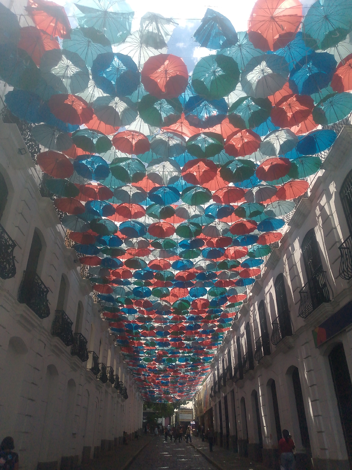 The image shows a street in Caracas, Venezuela, decorated with a dense canopy of colorful umbrellas. The umbrellas are primarily red, blue, and green and are suspended above the pedestrian street between two rows of buildings. The buildings are white with arched windows and wrought-iron balconies. People are walking along the street. The time of day appears to be midday, as the light is bright. The street is paved and relatively clean.