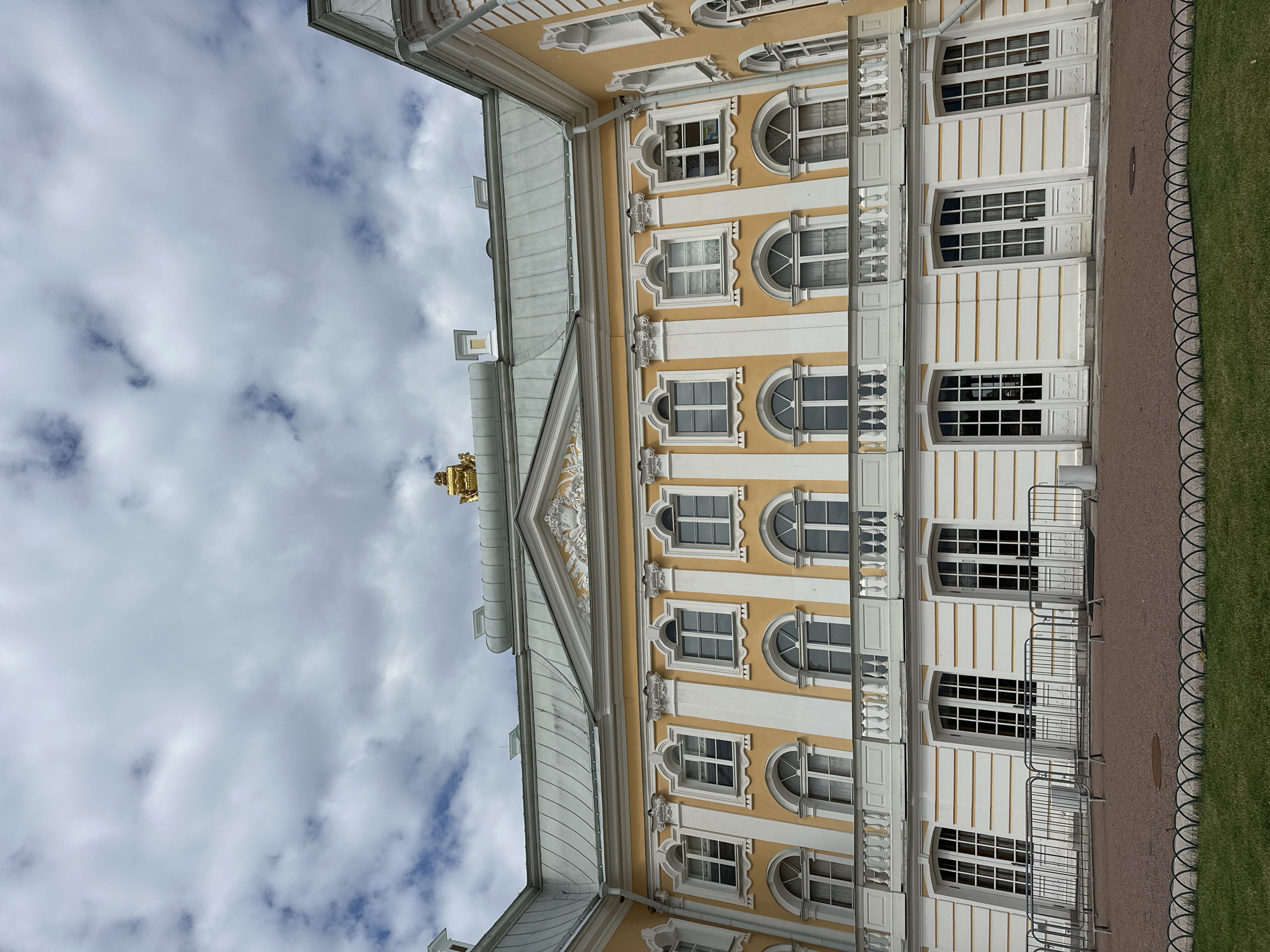 The image shows a facade of the Grand Palace in Peterhof, Russia. The palace is painted yellow with white architectural details and features a green roof. The facade has multiple windows and doors, many of which are arched. A balcony with a white railing runs along the second story. In front of the palace, there is a paved area with a low metal fence and a patch of green grass. The sky above is partly cloudy.